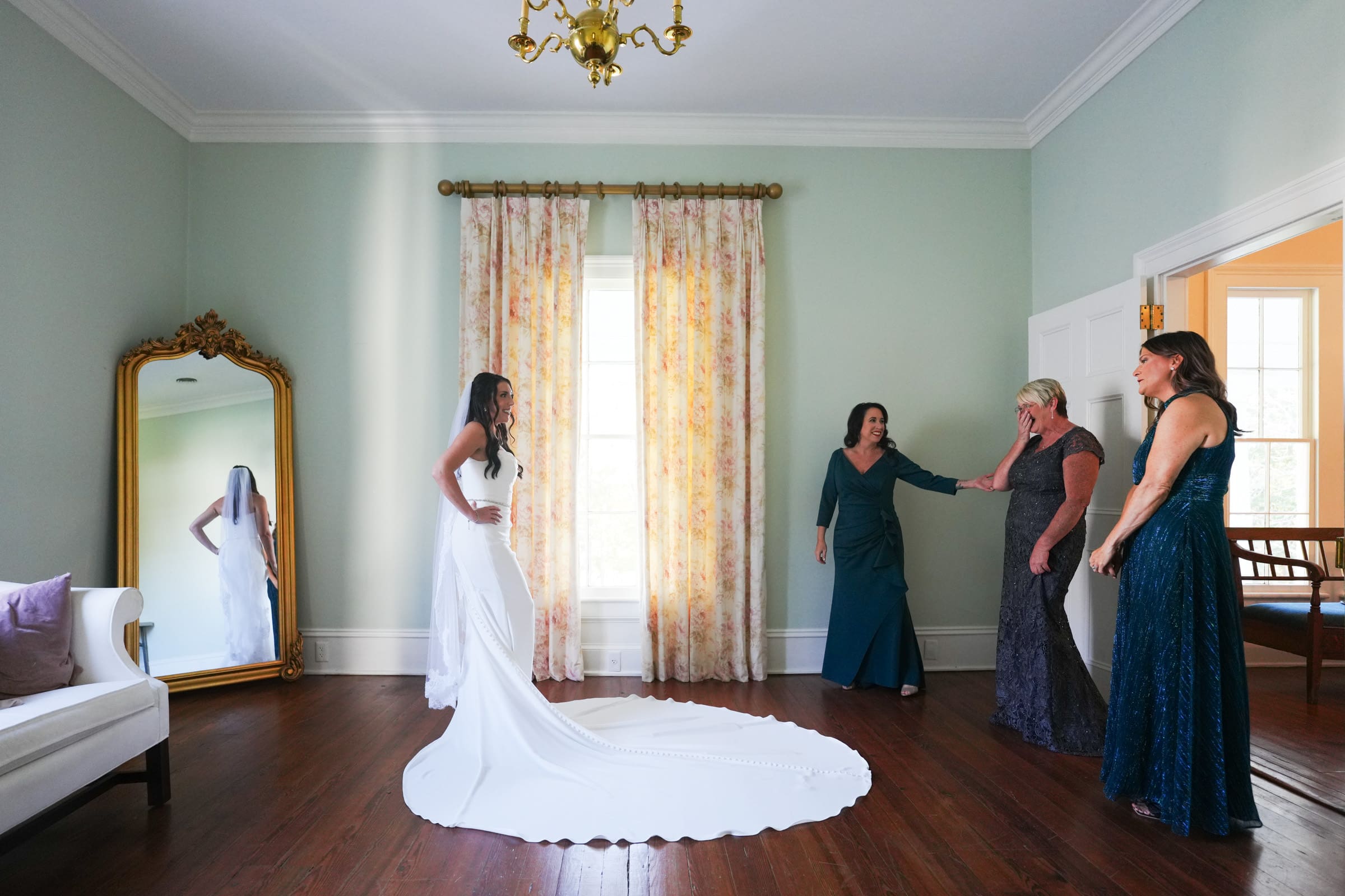 A bride in a white gown stands smiling before three women in colorful dresses at the Rose Hill Estate, near a large mirror reflecting her image. The room exudes vintage charm with wooden floors, pale green walls, a floral curtain, and a chandelier.