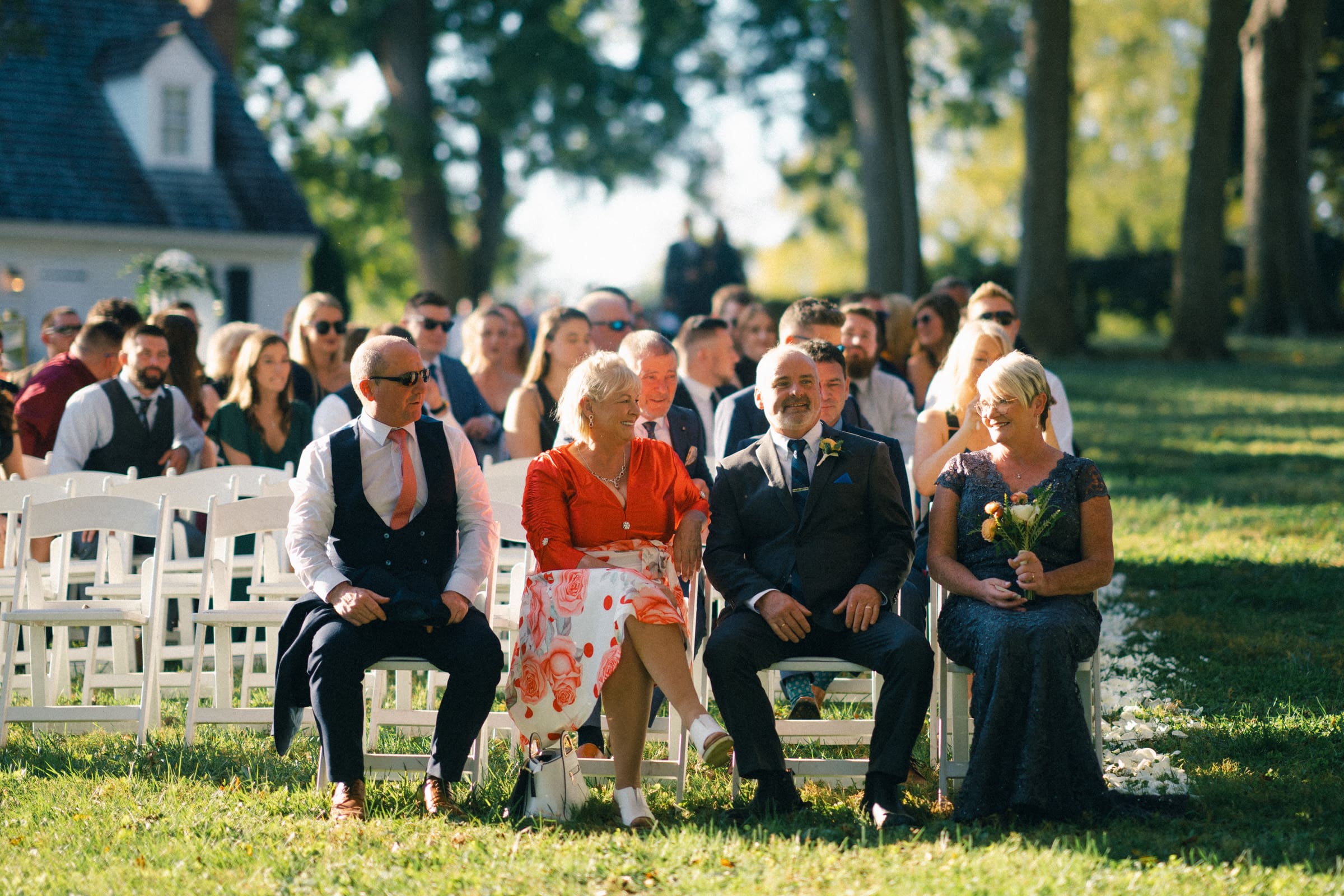 At a picturesque Rose Hill Estate wedding, a group of people sit on white chairs outdoors. The focus is on four individuals in the front row, smiling and dressed in formal attire. In the background, more guests are seated, surrounded by lush greenery and trees.