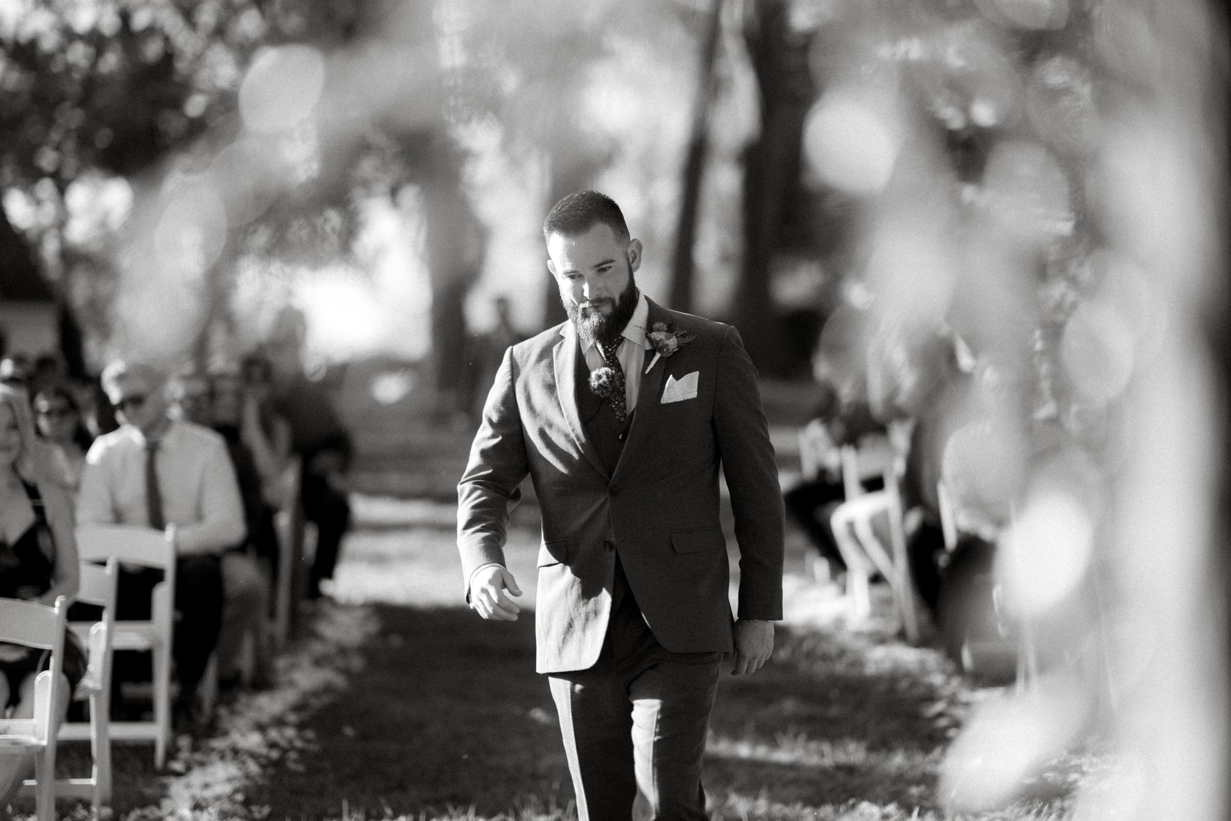 A black and white image captures a groom walking down an outdoor aisle at a Rose Hill Estate wedding. Wearing a suit with a boutonnière, he strides confidently past guests seated in neat rows. Sunlight gracefully filters through the surrounding trees, adding a touch of magic to the moment.