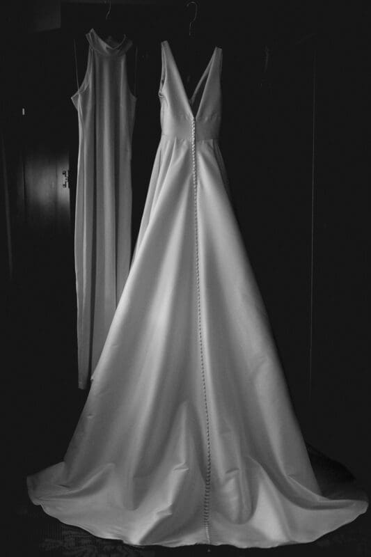 Carolina Inn Wedding - Frances & Lewis A black and white image captures the charm of a Carolina Inn wedding, with two long dresses elegantly hanging on hangers. The foreground dress features a sweeping train and buttons down the back, while a simpler sleeveless dress gently hangs in the background.