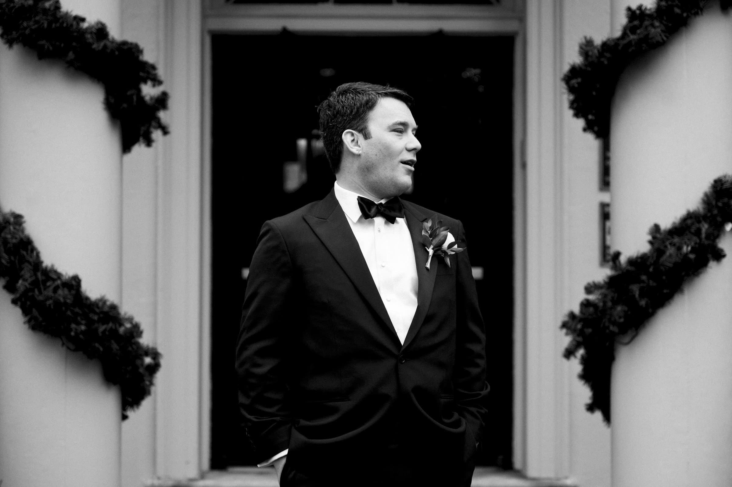 Carolina Inn Wedding - Frances & Lewis A man in a tuxedo with a bow tie stands at the entrance of the Carolina Inn, adorned with garlands. With hands in pockets, he gazes thoughtfully to the side in this elegant black-and-white image that captures the timeless essence of a wedding moment.