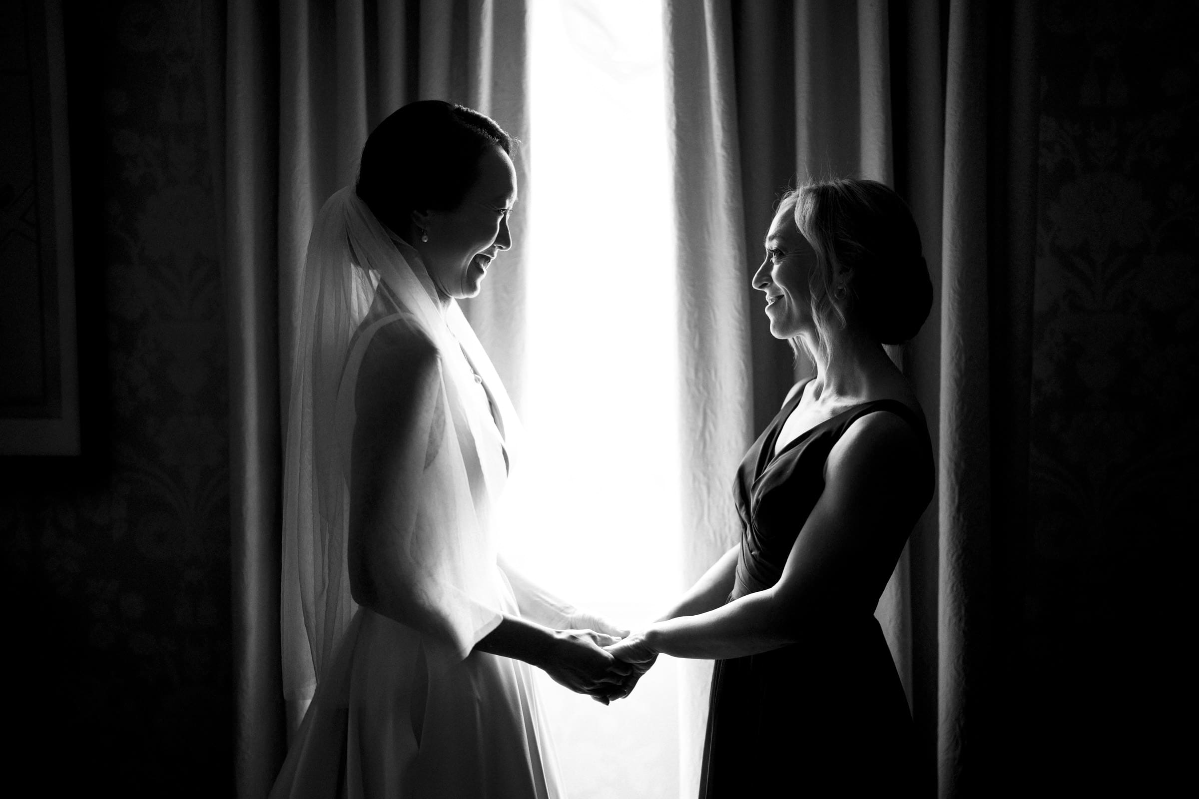 Carolina Inn Wedding - Frances & Lewis A black and white photograph captures two people standing before a sunlit window with delicate curtains at the Carolina Inn. The person on the left wears a wedding dress and veil, while the one on the right is dressed in a dark gown. They hold hands, smiling radiantly at each other.