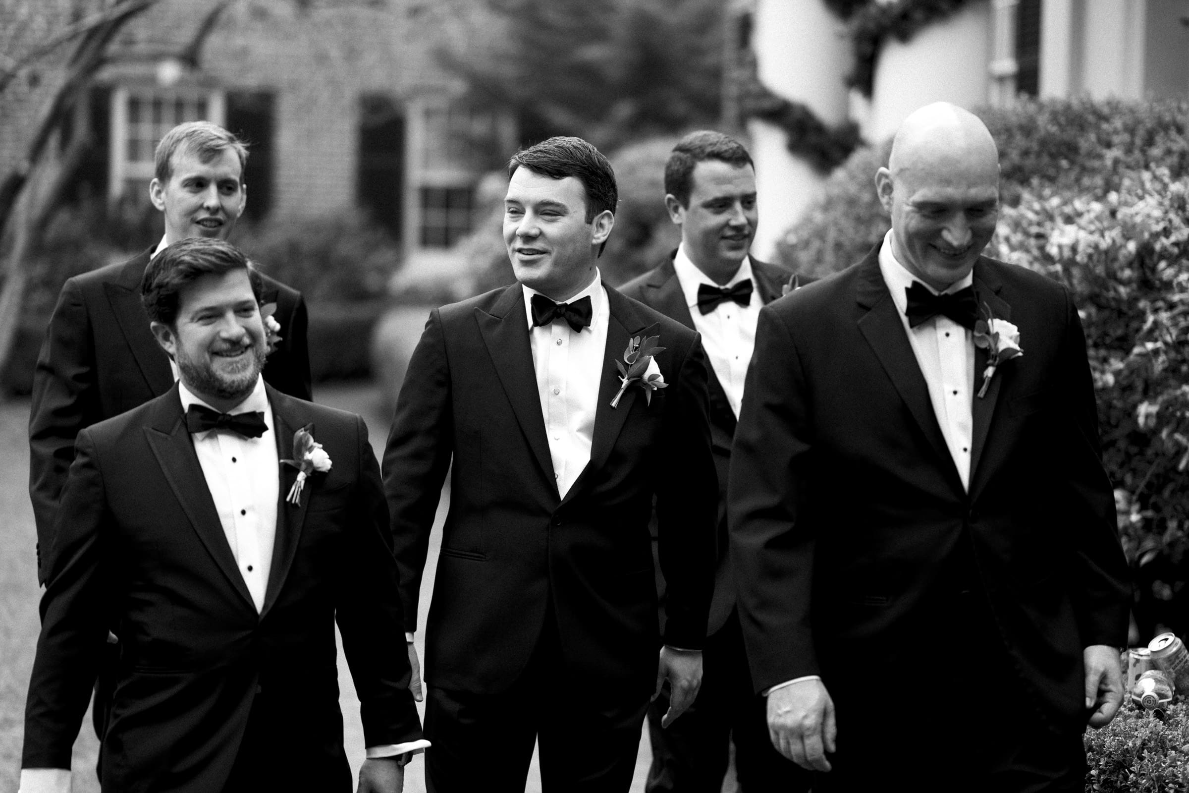 Carolina Inn Wedding - Frances & Lewis A group of five men in tuxedos with bow ties stroll outdoors, smiling and engaged in conversation. The backdrop of greenery and a quaint building hints at a Carolina Inn Wedding. This charming photo captures the moment in timeless black and white.
