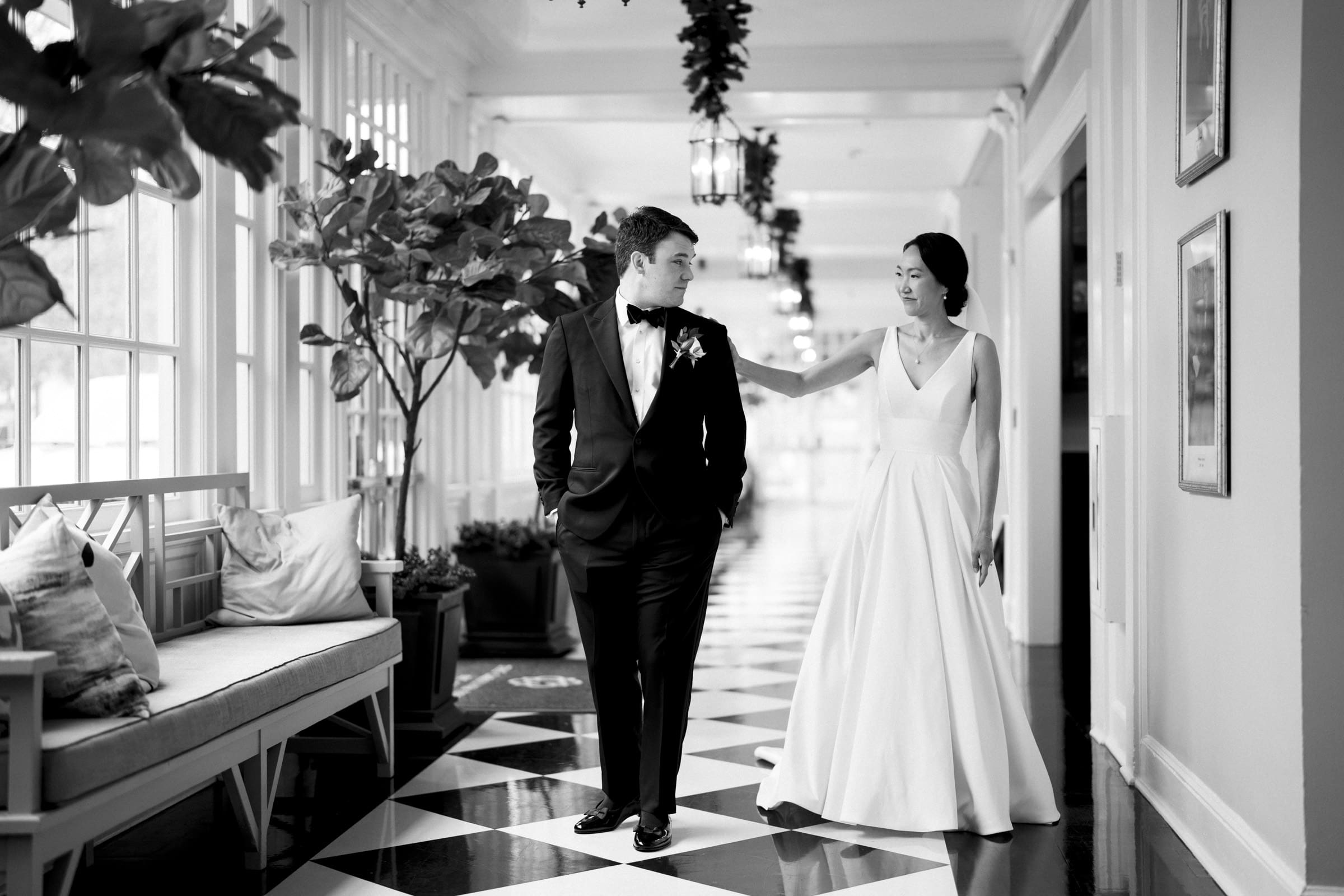Carolina Inn Wedding - Frances & Lewis A bride and groom walk hand in hand down a hallway with checkered flooring and large windows at the Carolina Inn. The bride wears a long, flowing gown, and the groom is in a tuxedo. They share a joyful moment, surrounded by elegant decor. Black and white image captures their timeless romance.
