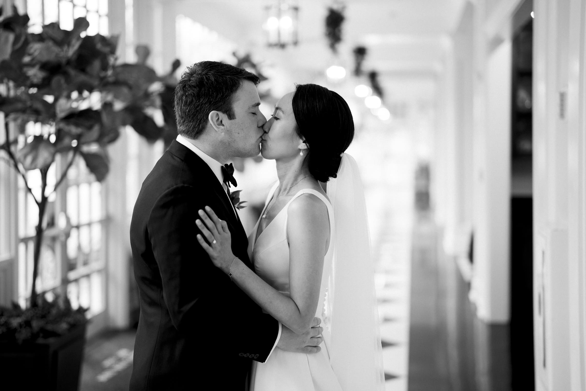 Carolina Inn Wedding - Frances & Lewis A bride and groom share a kiss in a hallway at the Carolina Inn, surrounded by lush plants. Dressed in formal wedding attire, with the bride elegantly veiled, the scene is captured in black and white, evoking a timeless feel that perfectly complements their cherished moment.