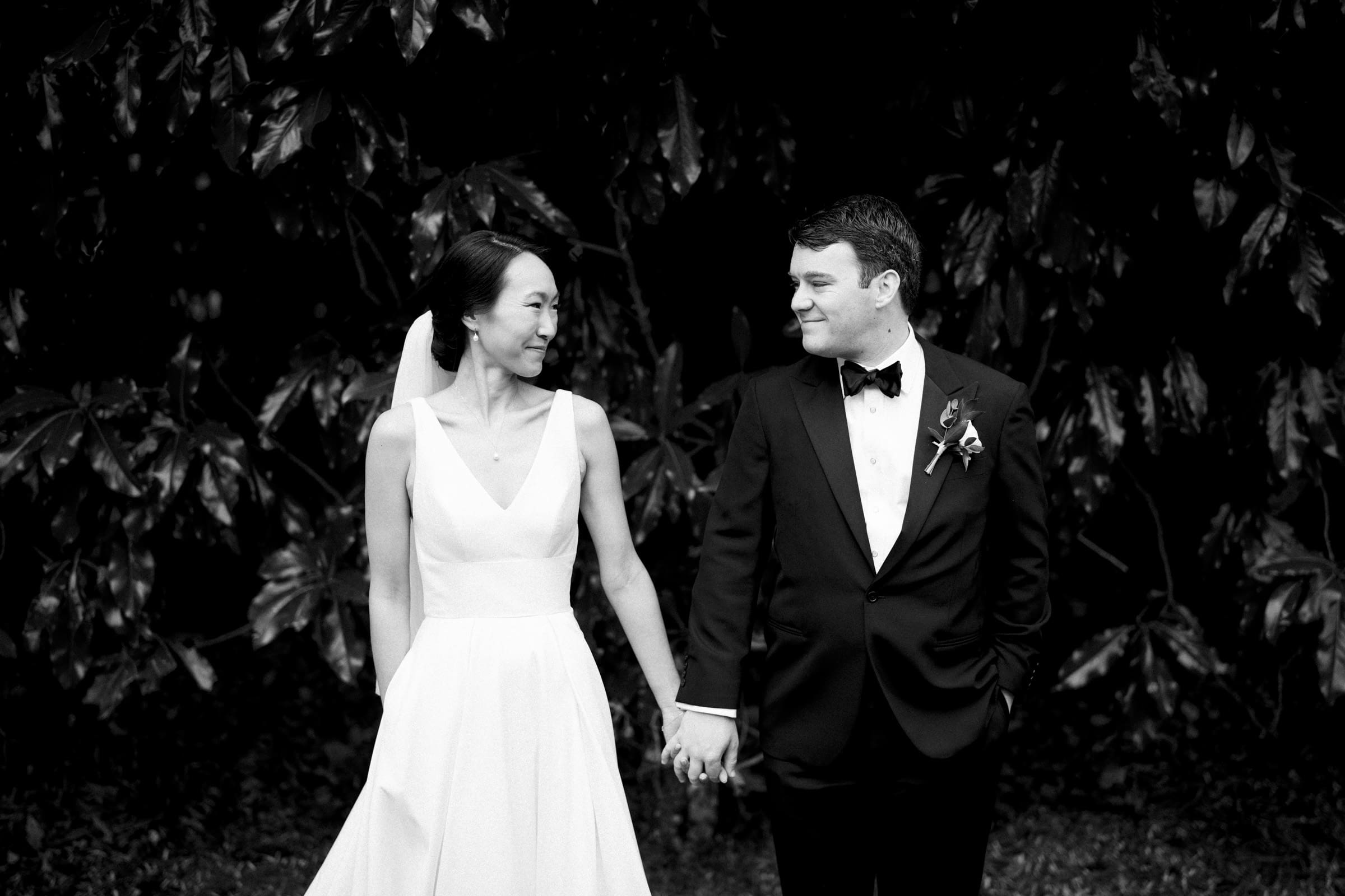 Carolina Inn Wedding - Frances & Lewis A bride and groom smile at each other while holding hands, captured in an enchanting black-and-white photo. The bride dazzles in her white dress and veil, the groom in a dark suit with a bow tie. They stand against lush foliage, embodying the timeless elegance of a Carolina Inn Wedding.