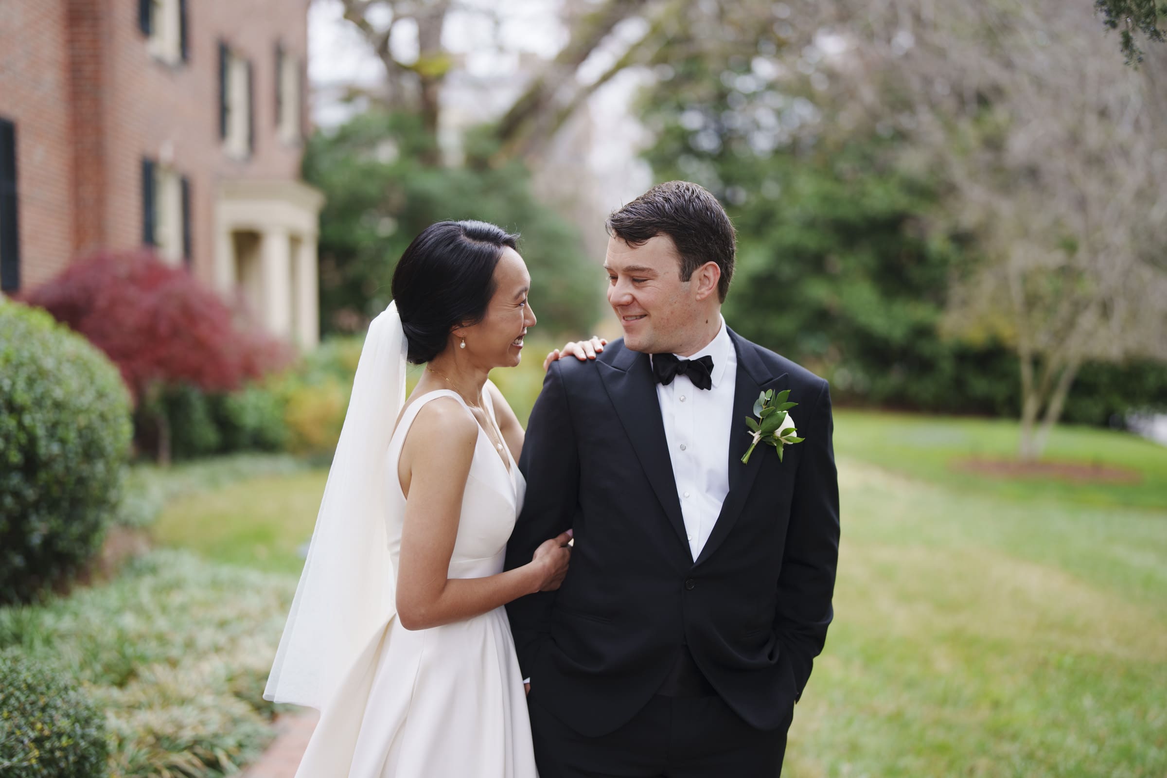 Carolina Inn Wedding - Frances & Lewis A bride and groom stand together, smiling outdoors at their Carolina Inn wedding. The bride, in a white dress and veil, gazes at the groom in his black tuxedo with a boutonniere. They are surrounded by lush greenery and the charming brick exterior of the inn.