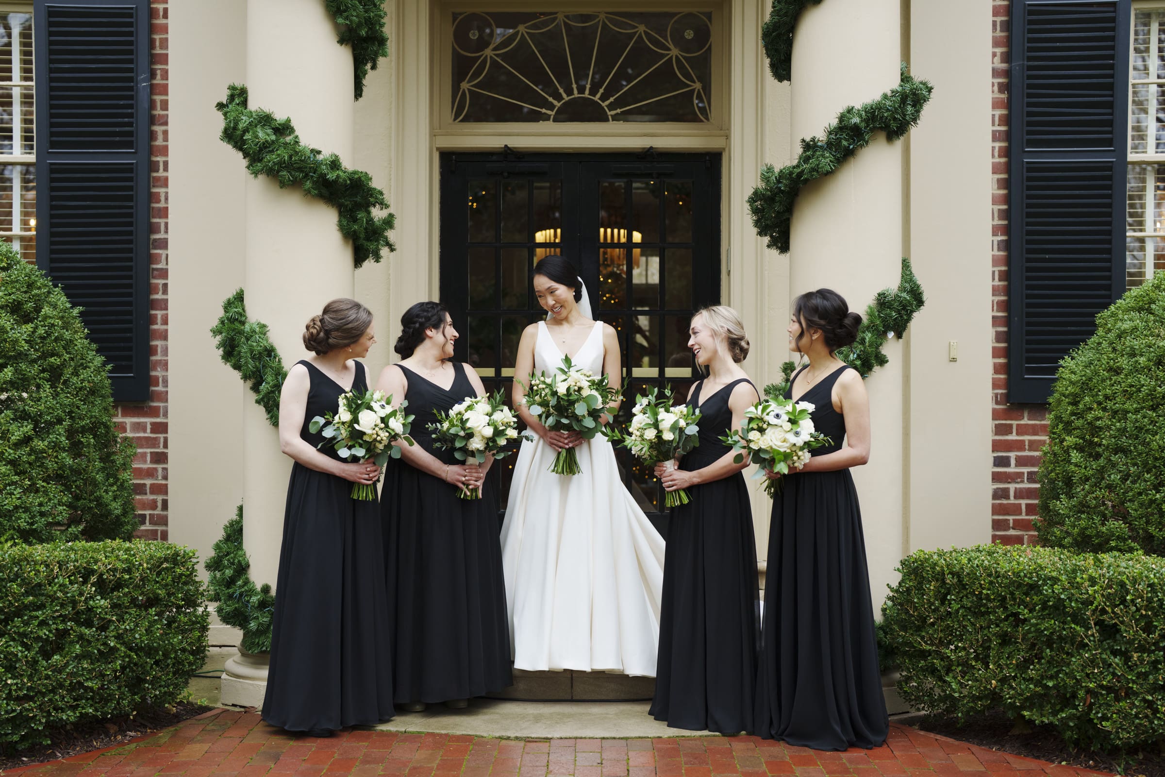 Carolina Inn Wedding - Frances & Lewis At the timeless Carolina Inn wedding, a bride in a white gown stands with four bridesmaids in black dresses holding bouquets, framed by manicured shrubs and brickwork. The elegant building adorned with green garlands sets a classic and sophisticated atmosphere.