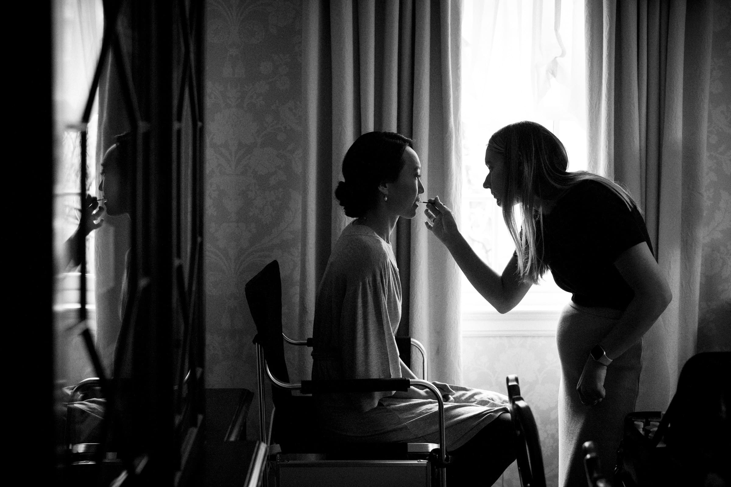 Carolina Inn Wedding - Frances & Lewis A black and white photo captures a woman at the Carolina Inn Wedding, sitting by a window as another applies her makeup. The room's patterned wallpaper and curtains add elegance, while the scene beautifully reflects in a nearby mirror.