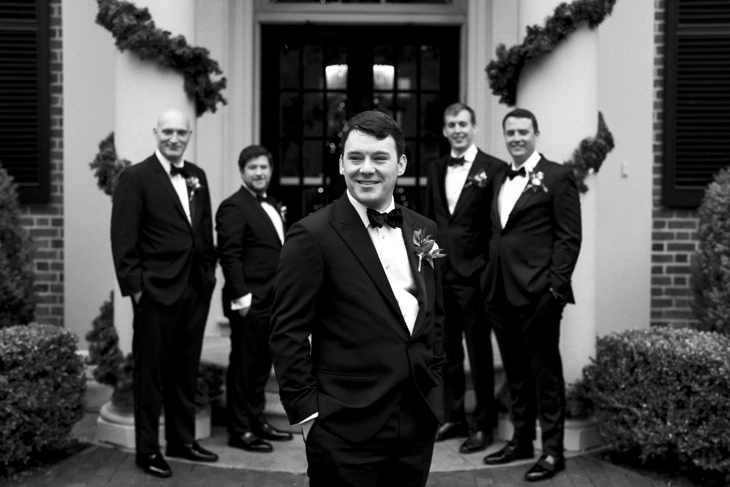 Carolina Inn Wedding - Frances & Lewis A groom stands with four groomsmen outside the Carolina Inn, all dressed in black tuxedos with bow ties and floral boutonnieres. The setting, adorned with greenery, exudes a formal elegance. This timeless black-and-white image captures the essence of a classic Carolina Inn wedding.