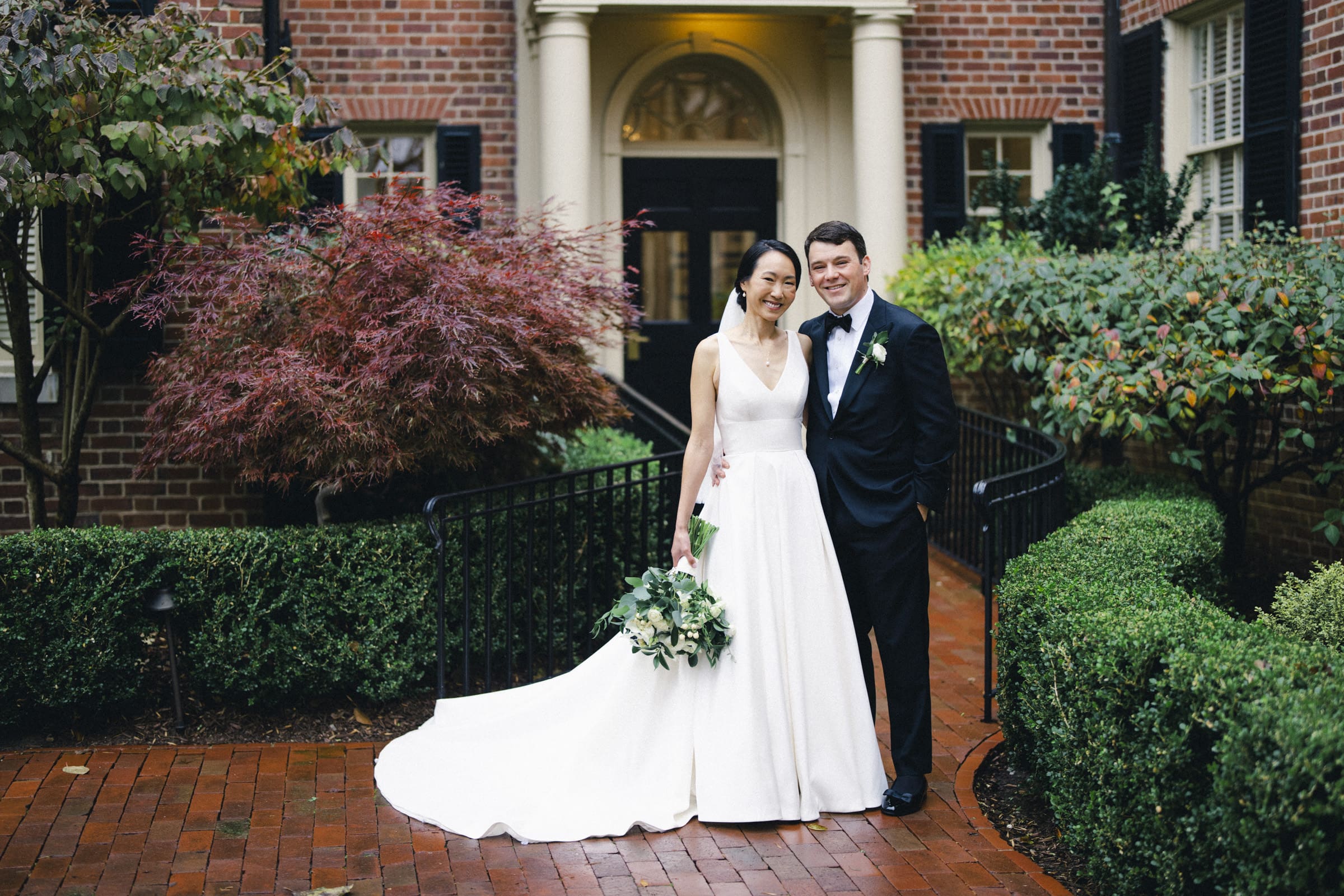Carolina Inn Wedding - Frances & Lewis A bride and groom stand smiling in a garden courtyard at the Carolina Inn. The bride wears a white gown and holds a bouquet of flowers, while the groom is dressed in a black tuxedo. They are surrounded by lush green shrubbery and elegant brick architecture.