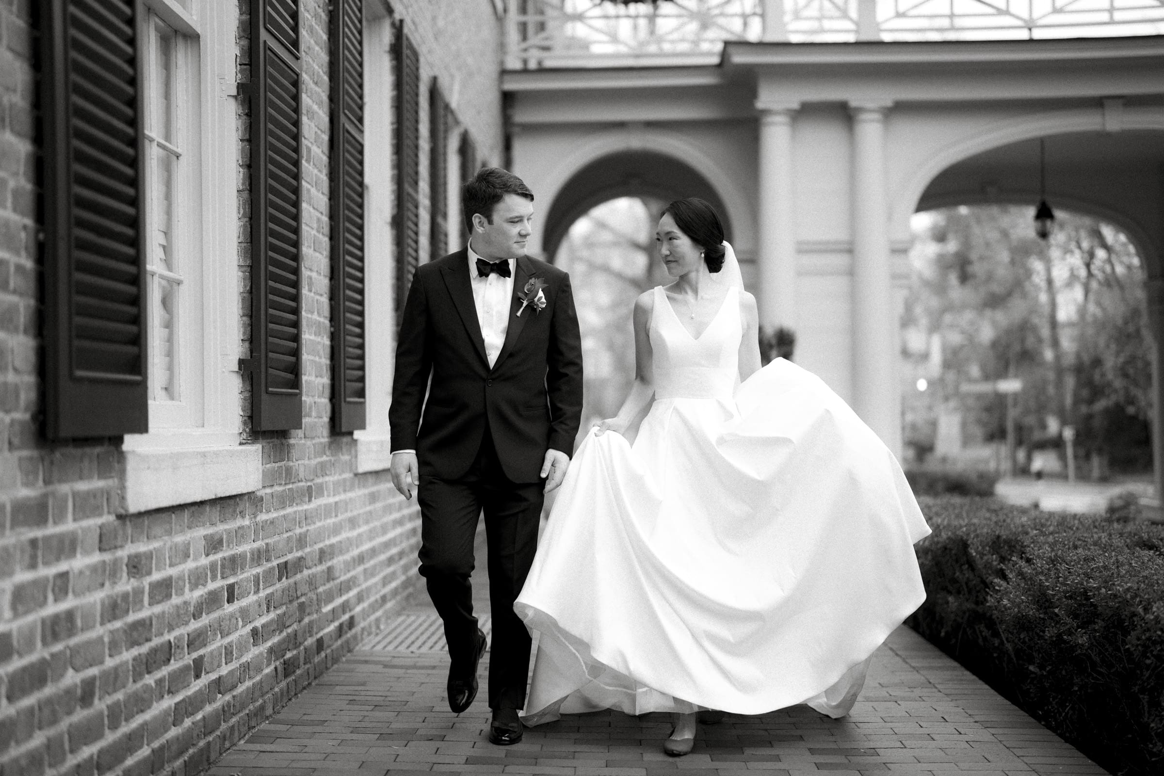 Carolina Inn Wedding - Frances & Lewis A black and white photo captures a couple in wedding attire strolling outside the Carolina Inn. The bride, in a long white gown, walks beside the groom, who is donned in a black suit with a bow tie, as they leisurely navigate the path by the brick building.