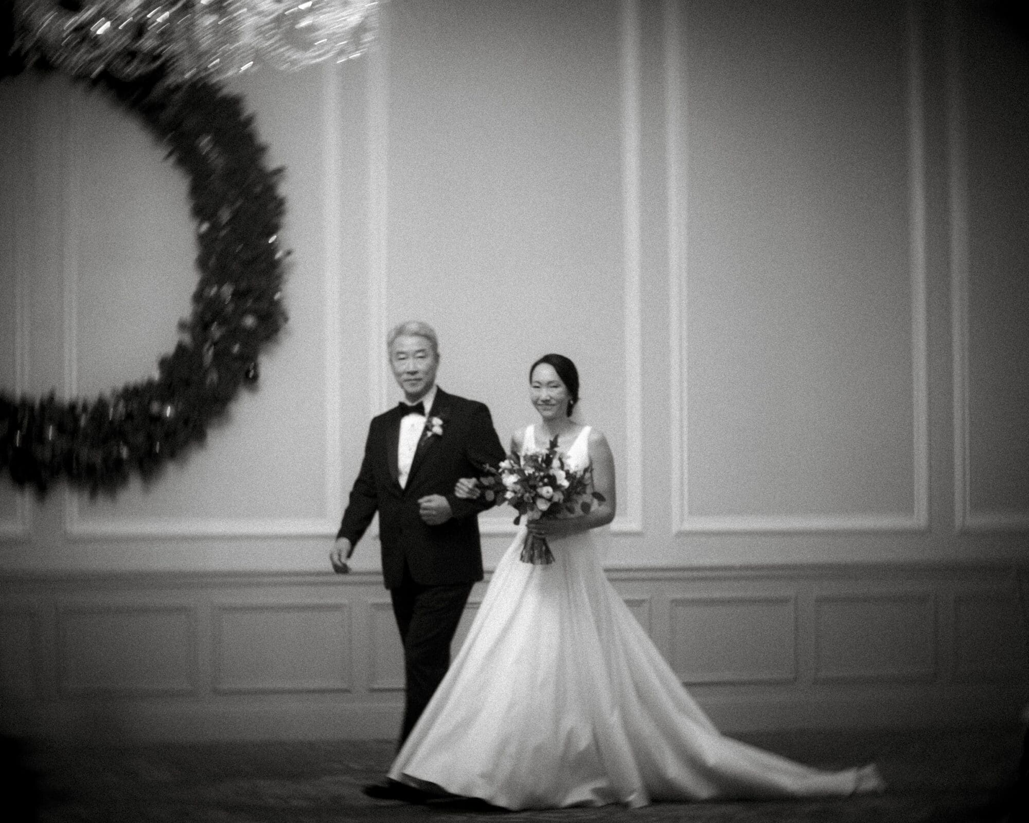 Carolina Inn Wedding - Frances & Lewis At the Carolina Inn Wedding, a bride in a flowing white dress walks down a hallway with an older man in a suit. She holds a bouquet and smiles. The black-and-white image captures the elegance, with a large decorative wreath adorning the wall.