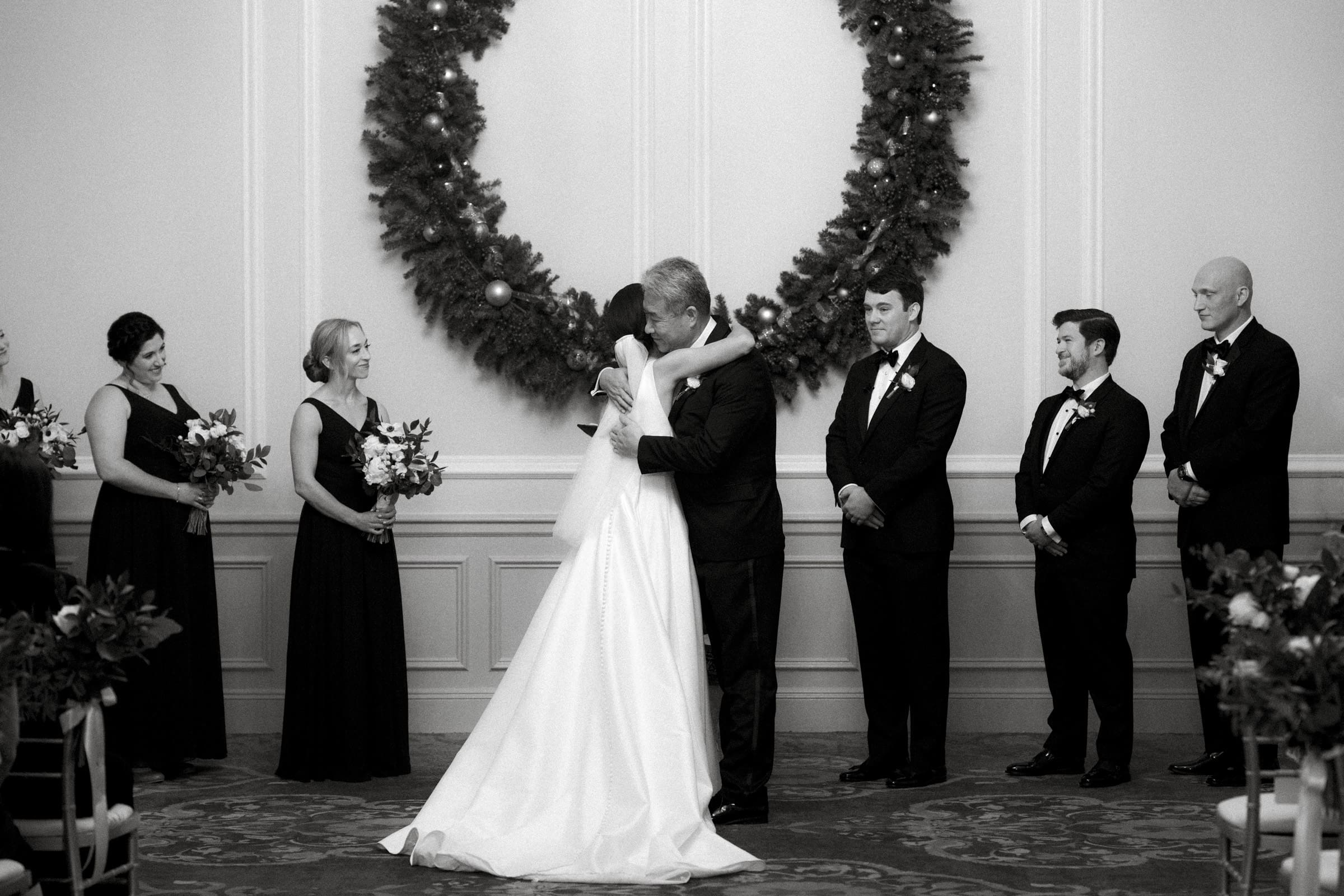 Carolina Inn Wedding - Frances & Lewis A bride and groom share a kiss in the elegant Carolina Inn setting, a large wreath behind them. Bridesmaids in black dresses hold bouquets on the left, while groomsmen in suits stand on the right, witnessing the couple's embrace.