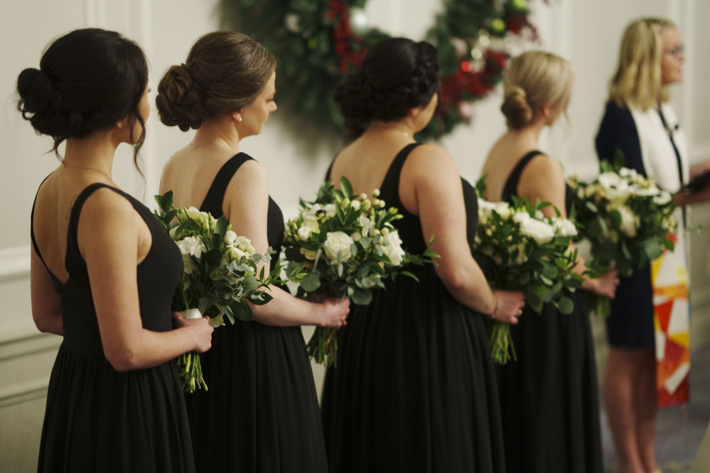 Carolina Inn Wedding - Frances & Lewis Four bridesmaids in black dresses hold white and green floral bouquets at a Carolina Inn wedding. They stand in a line facing the officiant, with a wreath visible in the background.