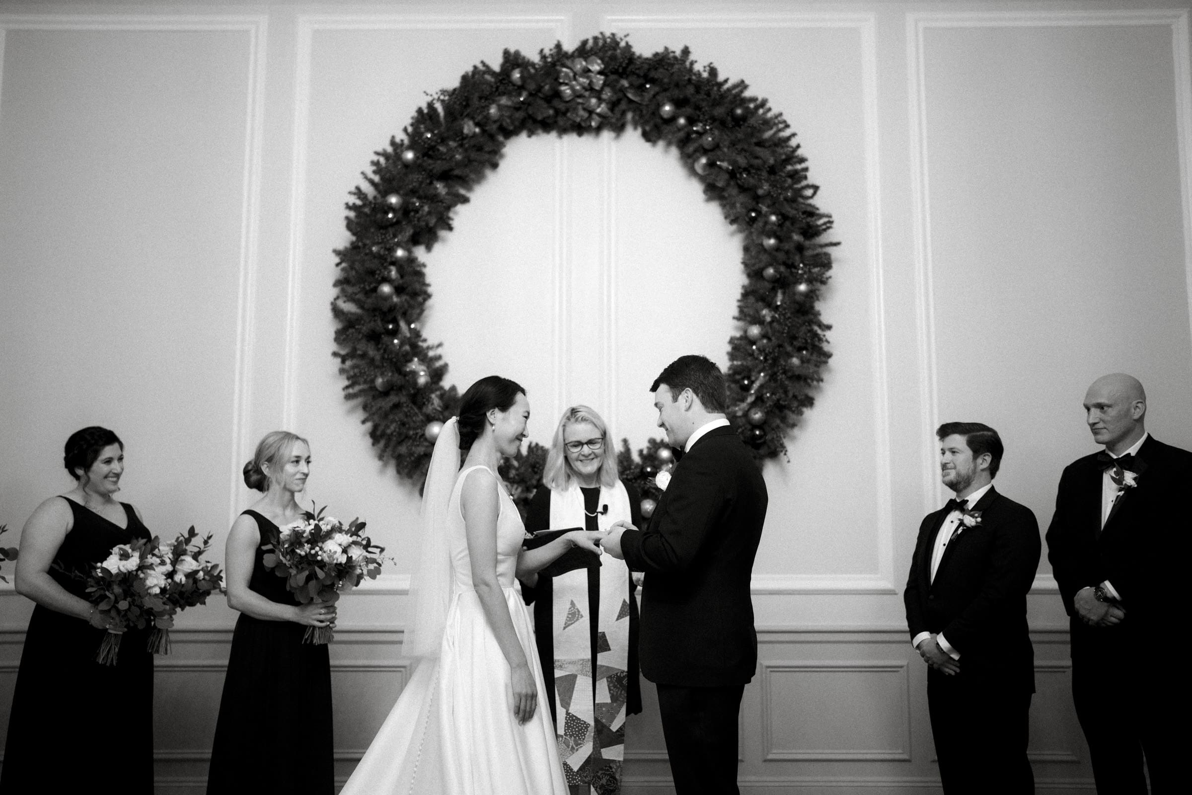 Carolina Inn Wedding - Frances & Lewis A Carolina Inn Wedding unfolds beautifully, with a couple exchanging vows beneath a large wreath. The bride dazzles in a white dress, the groom in his dark suit. Bridesmaids and groomsmen elegantly line the sides, bouquets in hand, adding to the formal grace of this memorable occasion.