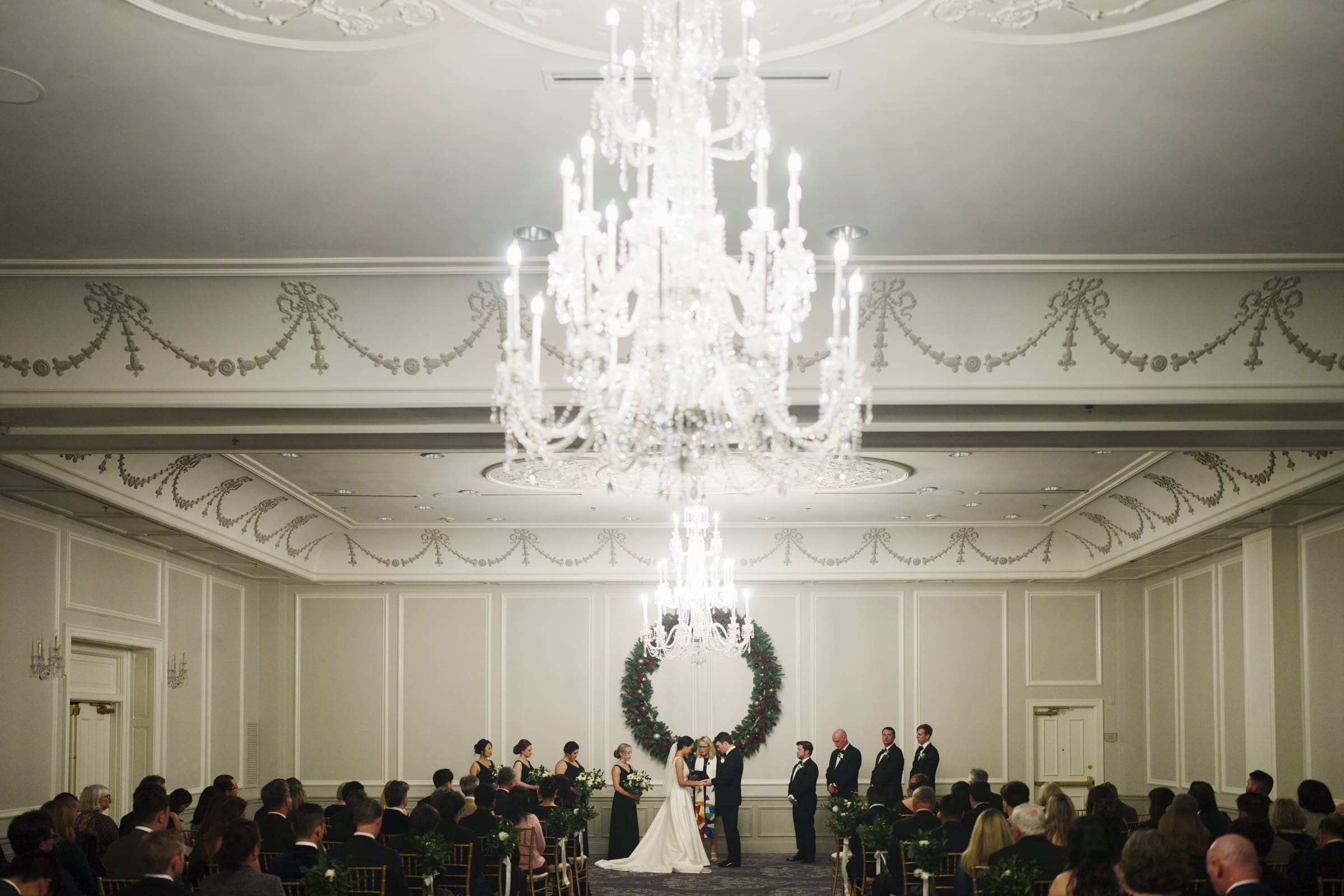 Carolina Inn Wedding - Frances & Lewis A Carolina Inn Wedding unfolds in an elegant ballroom with a grand chandelier. The bride and groom grace a floral archway, surrounded by their bridal party. Guests are seated in rows on either side, deeply engaged in the beautiful moment.