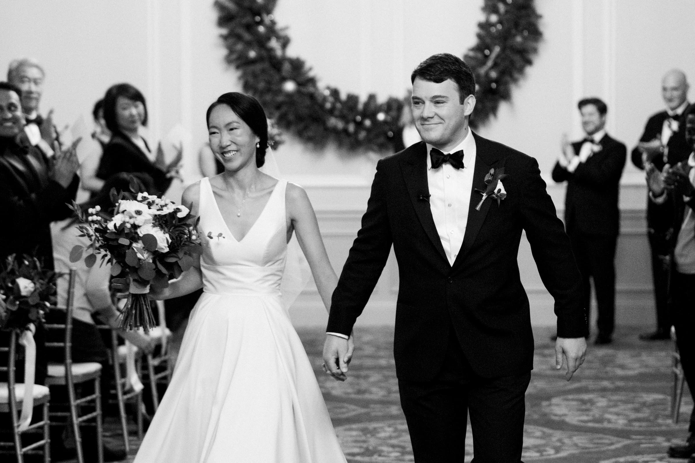 Carolina Inn Wedding - Frances & Lewis At a Carolina Inn wedding, a joyful couple, dressed in elegant attire, holds hands and walks down the aisle. The bride carries a bouquet while the groom sports a bow tie. Guests, smiling and clapping, are seated on either side as a large wreath hangs behind them.
