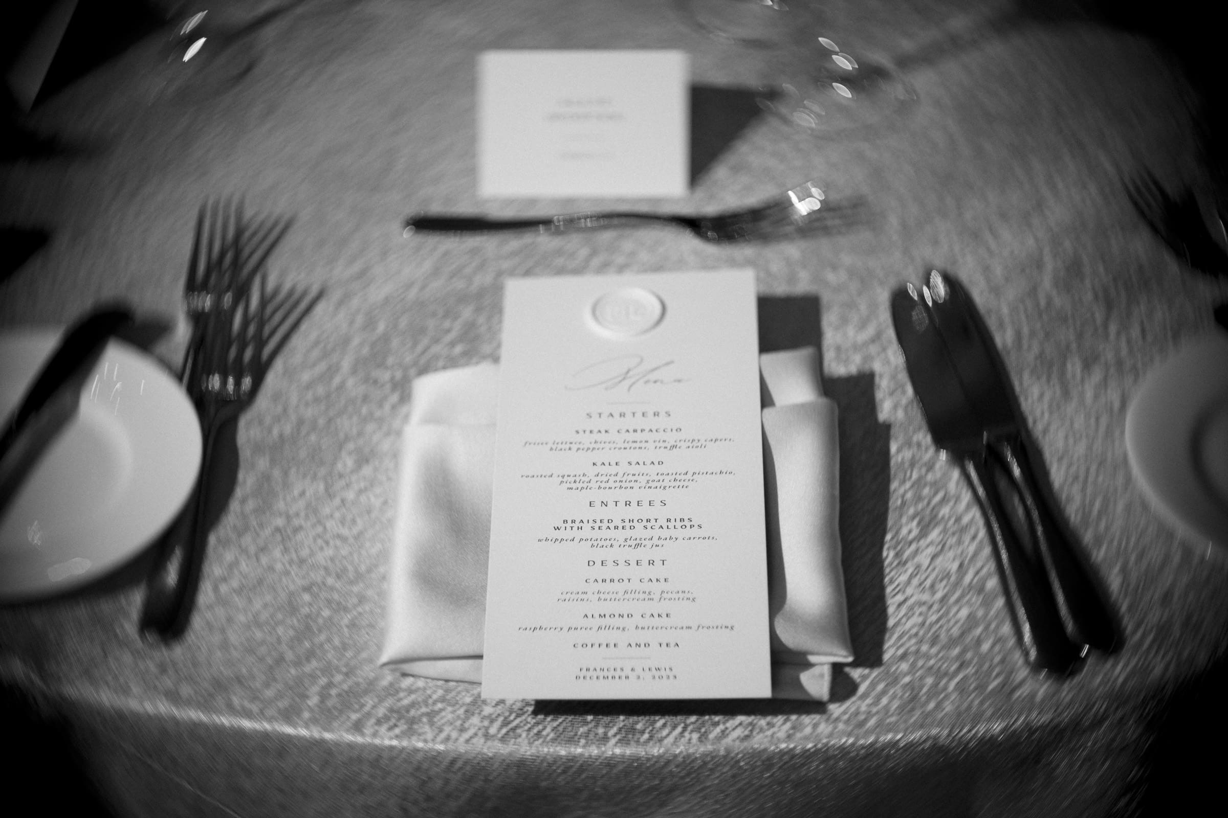 Carolina Inn Wedding - Frances & Lewis A black and white image showcasing an elegant Carolina Inn wedding table setting, with a menu placed on a folded napkin, surrounded by cutlery. The textured tablecloth adds depth, while the out-of-focus place card in the background hints at a day to remember.