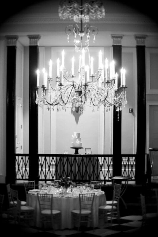 Carolina Inn Wedding - Frances & Lewis A black-and-white image of an elegant dining room at the Carolina Inn, featuring a large chandelier above a round table set for dinner. In the background, a tiered cake is displayed on a pedestal, adding to the formal setting's ornate decor—a picture-perfect venue for a wedding.
