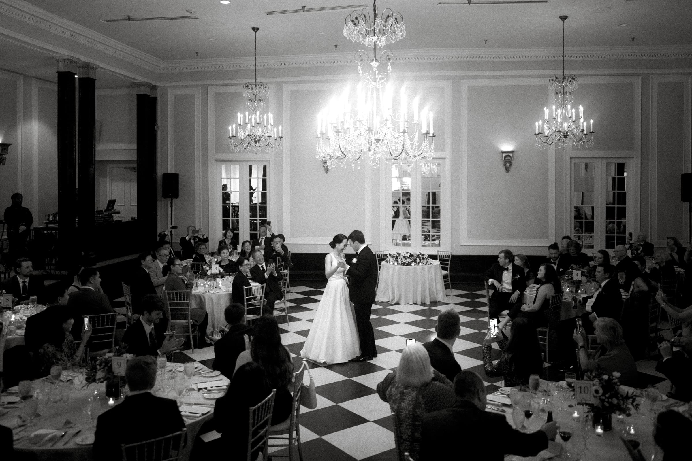 Carolina Inn Wedding - Frances & Lewis A black-and-white photo captures a couple sharing their first dance at a Carolina Inn Wedding. They are surrounded by seated guests in an elegant, chandelier-lit ballroom with a checkered floor.