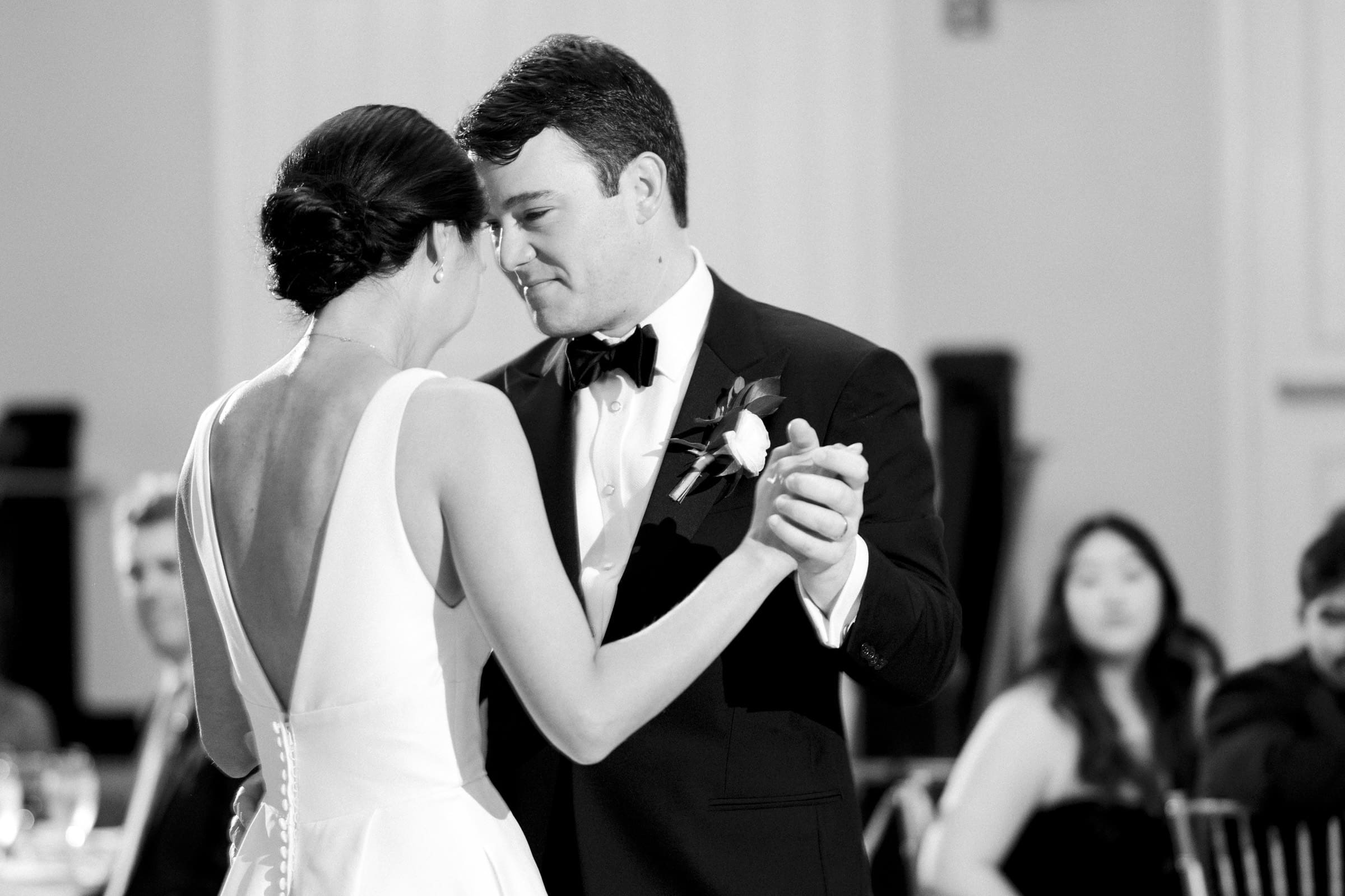 Carolina Inn Wedding - Frances & Lewis At a Carolina Inn wedding, a couple in formal attire shares a heartfelt dance. The bride stuns in her low-back white dress, while the groom dons a black suit with bow tie and boutonniere. They gaze into each other’s eyes amidst their seated guests.