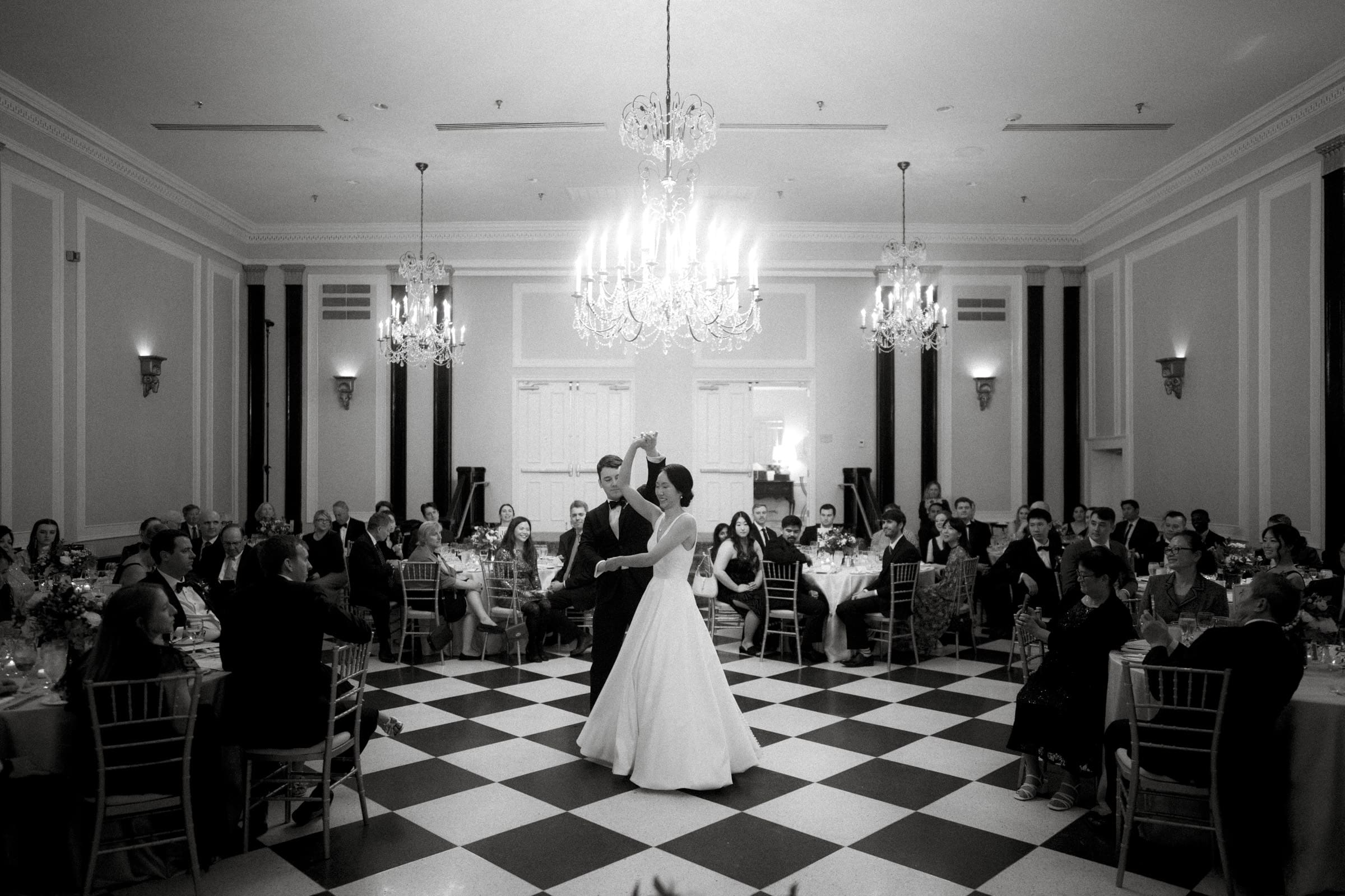 Carolina Inn Wedding - Frances & Lewis At a Carolina Inn wedding, a bride and groom share their first dance on a checkered floor in an elegant ballroom. Guests are seated around the room, watching the couple sway under a grand chandelier, enveloped in a classic black-and-white aesthetic.