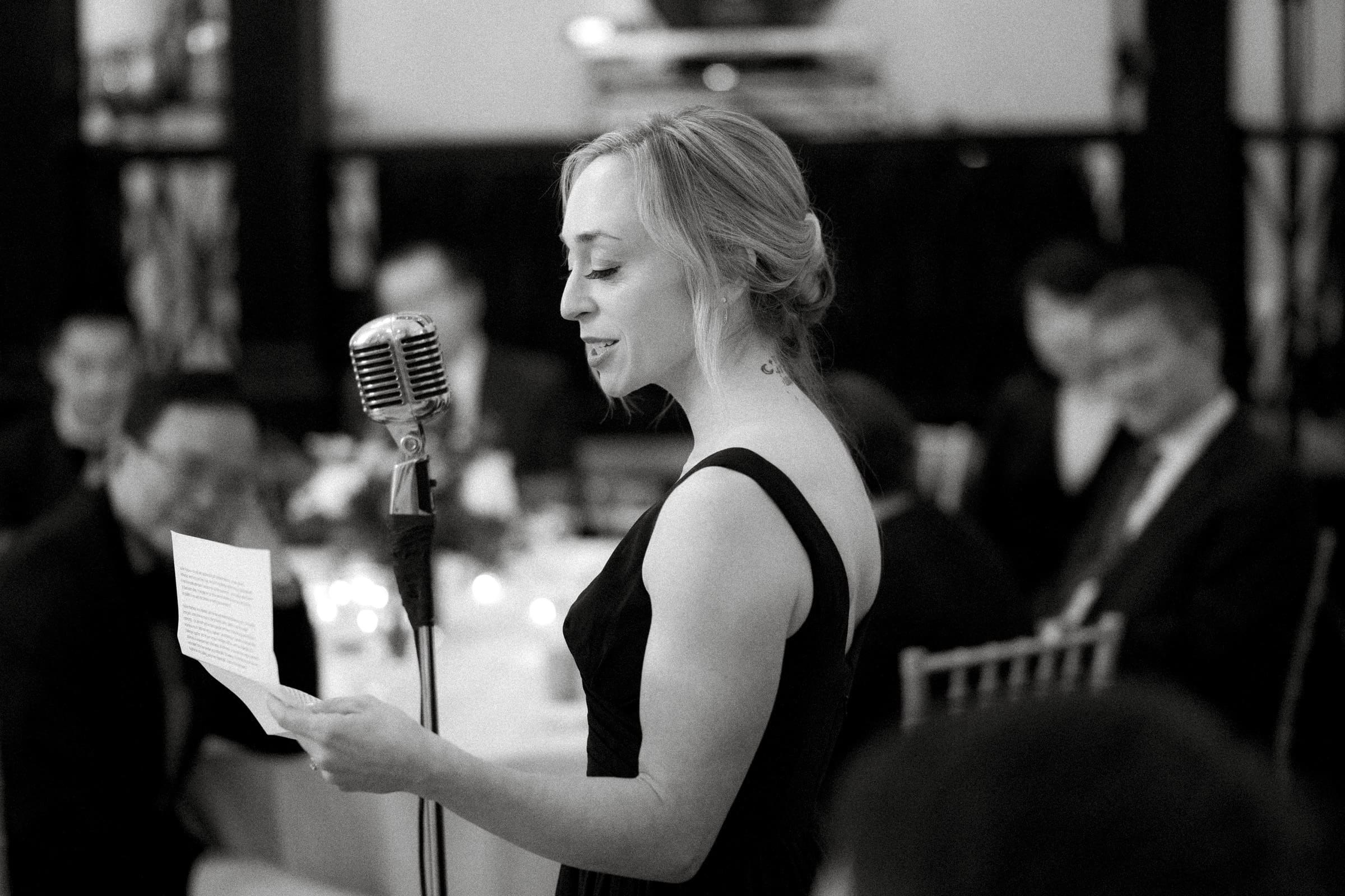 Carolina Inn Wedding - Frances & Lewis In a dimly lit room at the Carolina Inn, a woman in a formal dress speaks into a vintage microphone, holding a piece of paper. Seated guests linger in the background at what appears to be an elegant wedding gathering. The black and white image captures an air of timeless sophistication.