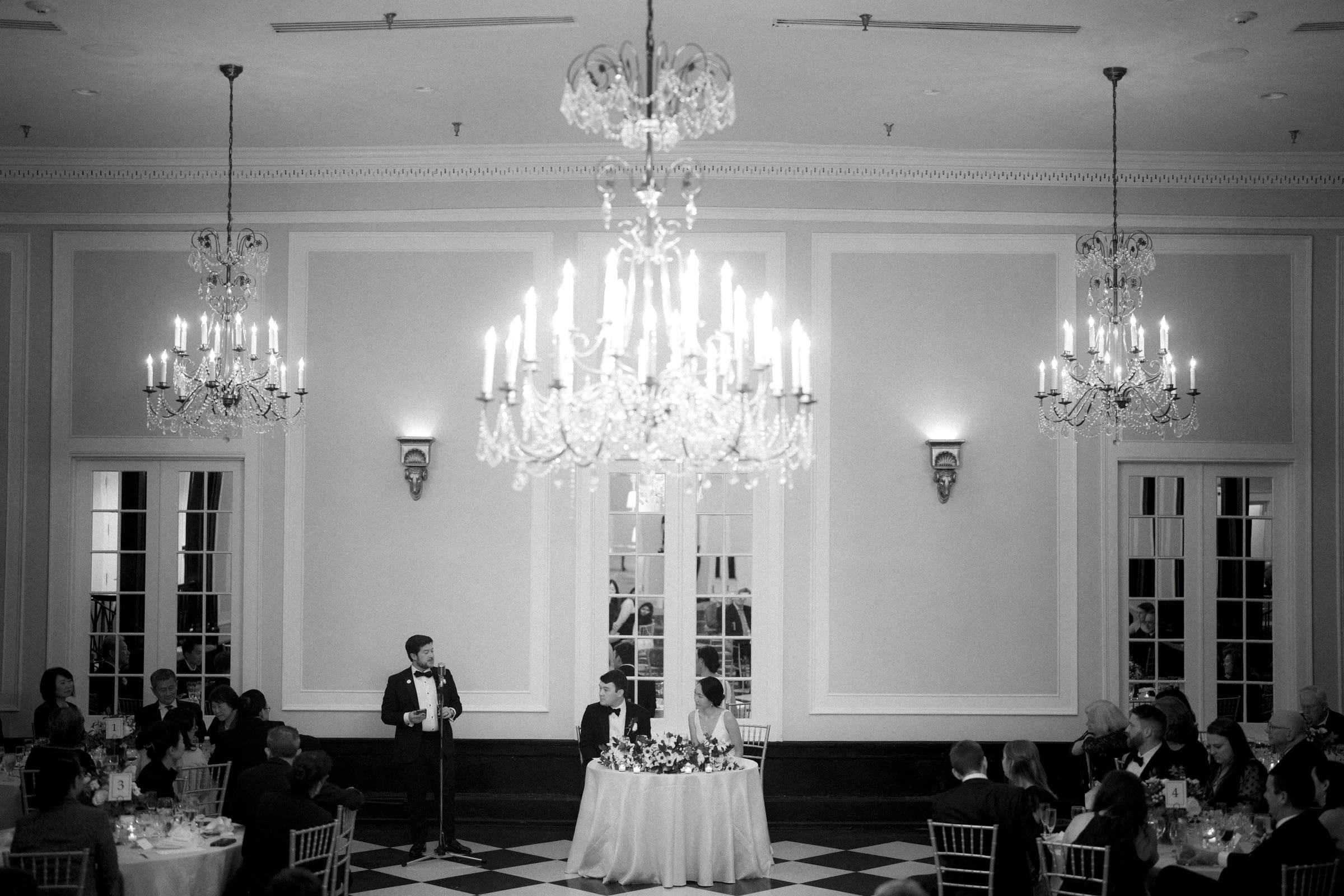 Carolina Inn Wedding - Frances & Lewis A black-and-white photo captures a formal event at the Carolina Inn, where chandeliers glisten above a man in a suit speaking into a microphone. He stands by a table with two seated guests, possibly celebrating their wedding, amidst round tables of attendees on the checkered floor.