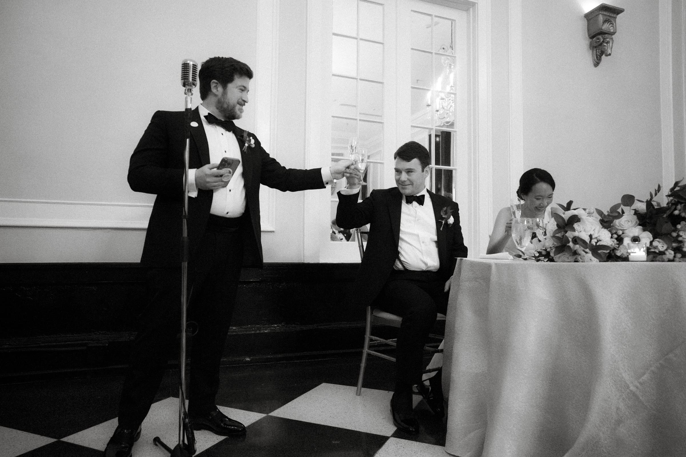 Carolina Inn Wedding - Frances & Lewis A black and white photo captures a man in a tuxedo raising a glass in a toast at what seems to be a Carolina Inn wedding. Another man, also in a tuxedo, sits at the table beside a woman in a dress, with a microphone nearby, highlighting the elegance of this formal event.