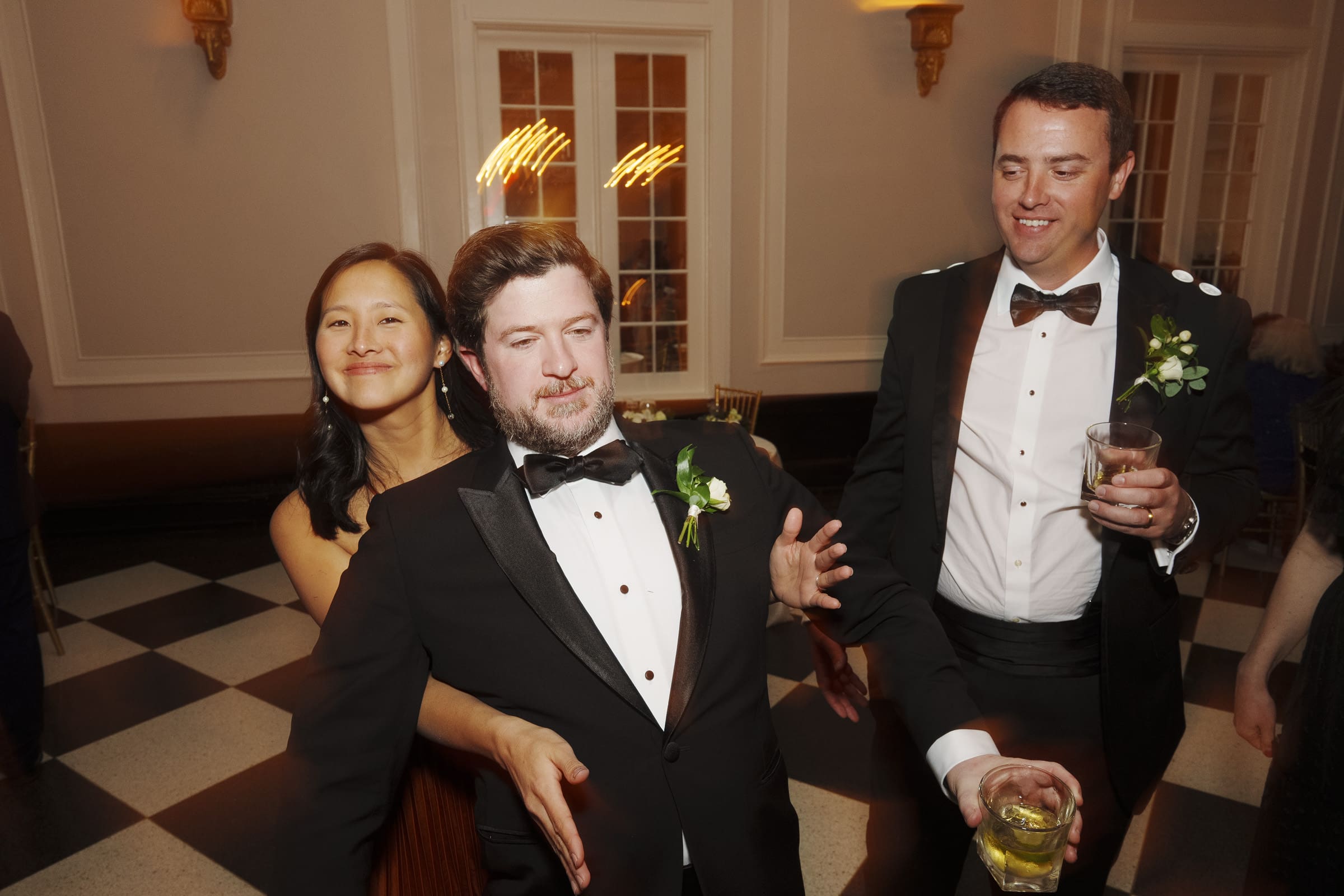 Carolina Inn Wedding - Frances & Lewis Three people in formal attire dance and smile at an elegant Carolina Inn wedding. Two men in black tuxedos with bow ties and a woman in a dress stand closely, each holding drinks, enjoying the festive moment on a checkered floor.