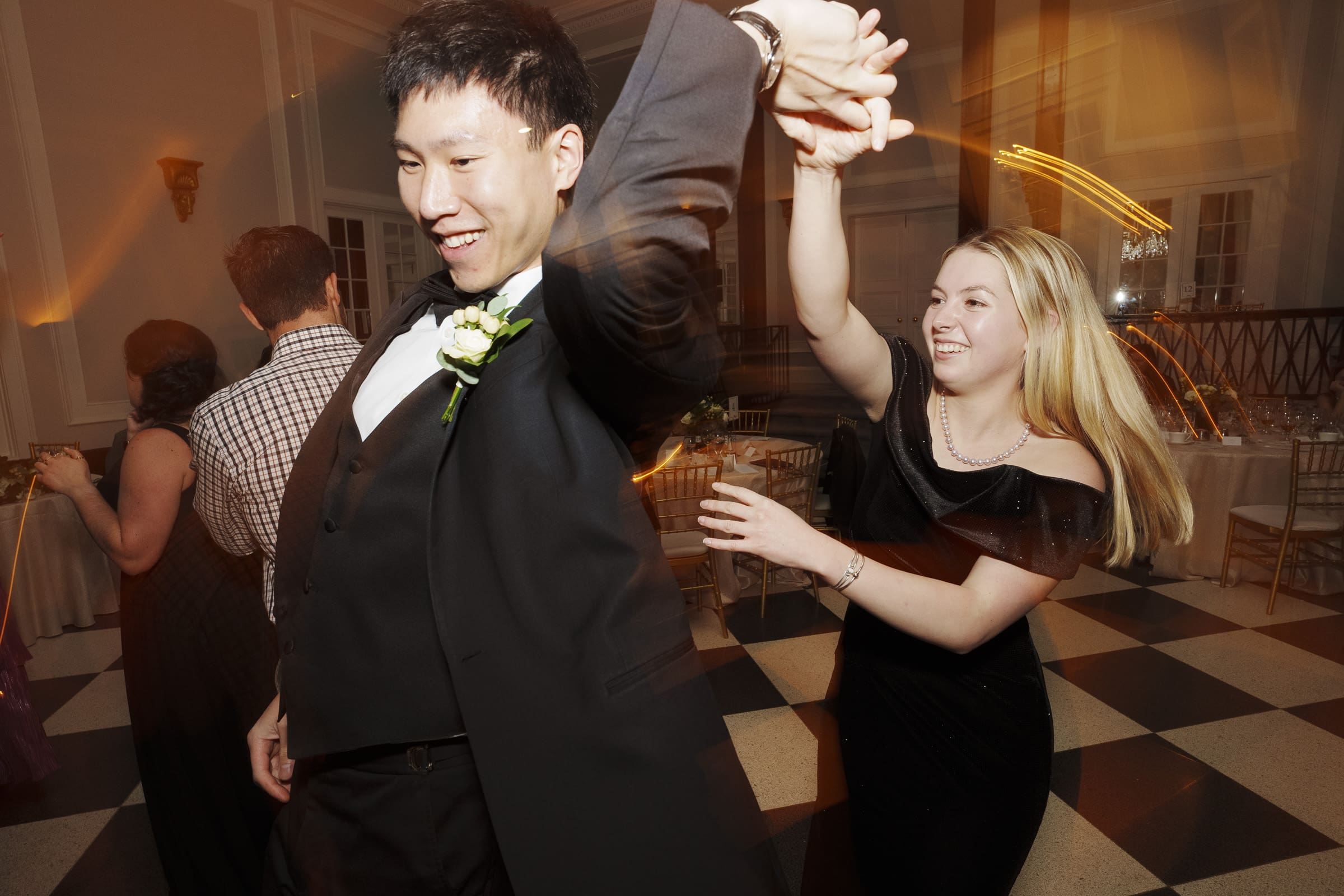 Carolina Inn Wedding - Frances & Lewis At a Carolina Inn wedding, a man and woman in formal attire dance joyfully. The man smiles broadly as he twirls the blonde-haired woman, both beaming with happiness. In the softly lit background, guests enjoy the elegant ambiance and festive atmosphere.