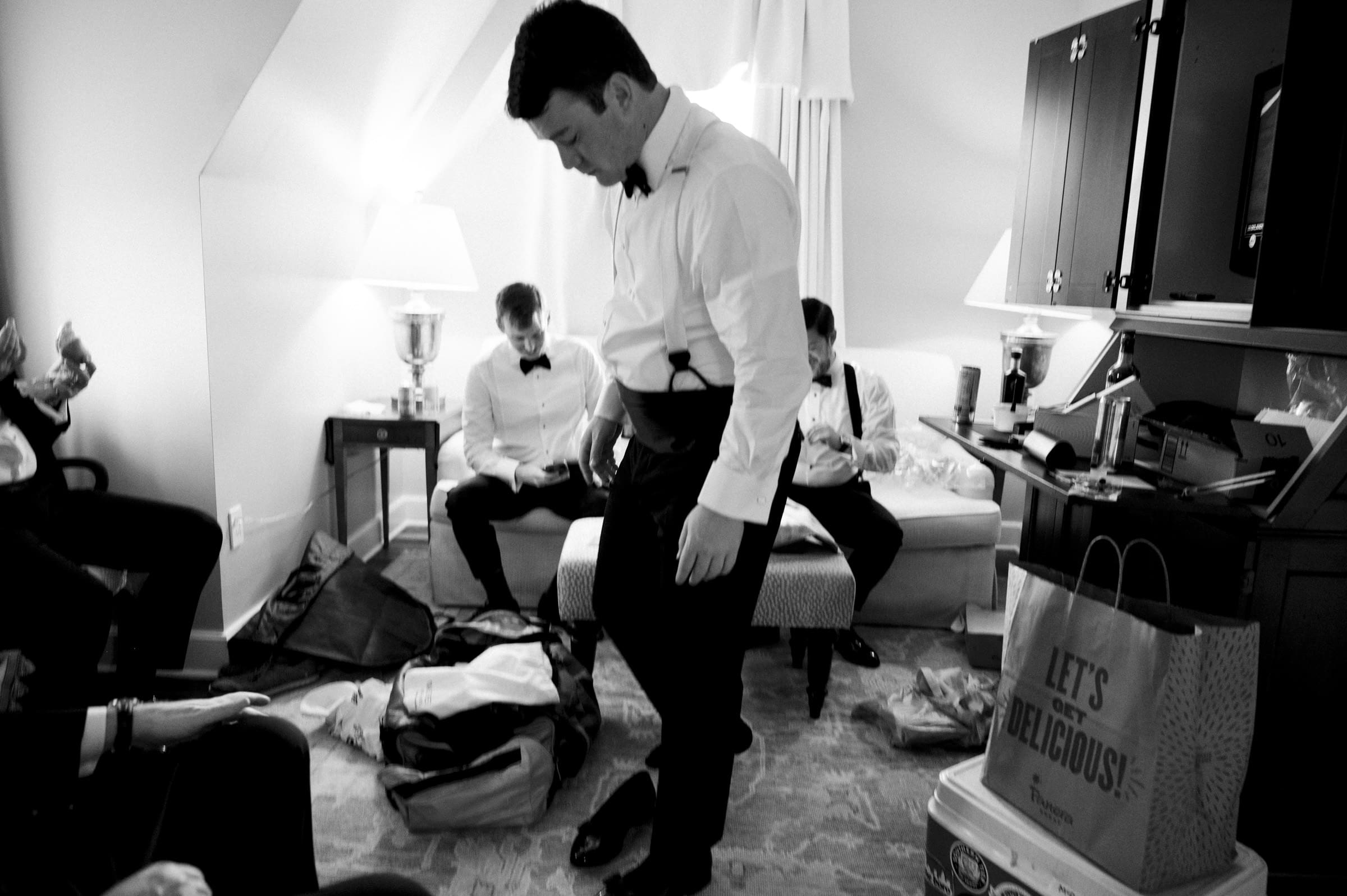 Carolina Inn Wedding - Frances & Lewis A black and white image captures the chaotic preparation for a Carolina Inn wedding. Four men are dressing in a cluttered room; the central figure adjusts his pants, while two others lounge on a couch and another sits on a chair. Bags, including a shopping bag, are scattered under the lamp's warm glow.