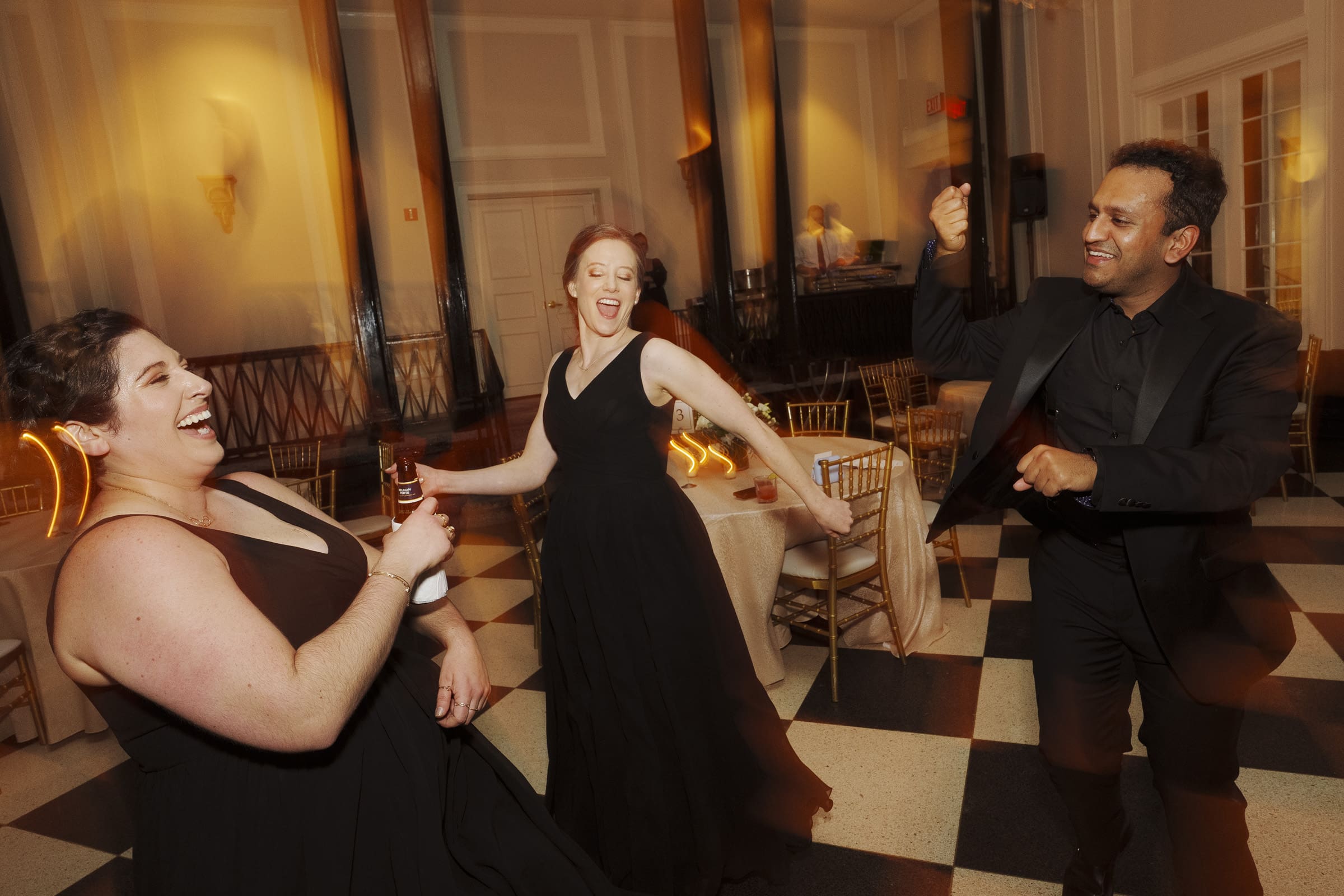Carolina Inn Wedding - Frances & Lewis Three people in formal black attire dance joyfully in a warmly lit room with checkered flooring, exuding the lively atmosphere of a Carolina Inn wedding celebration. One woman holds a drink, while the others smile and move energetically.