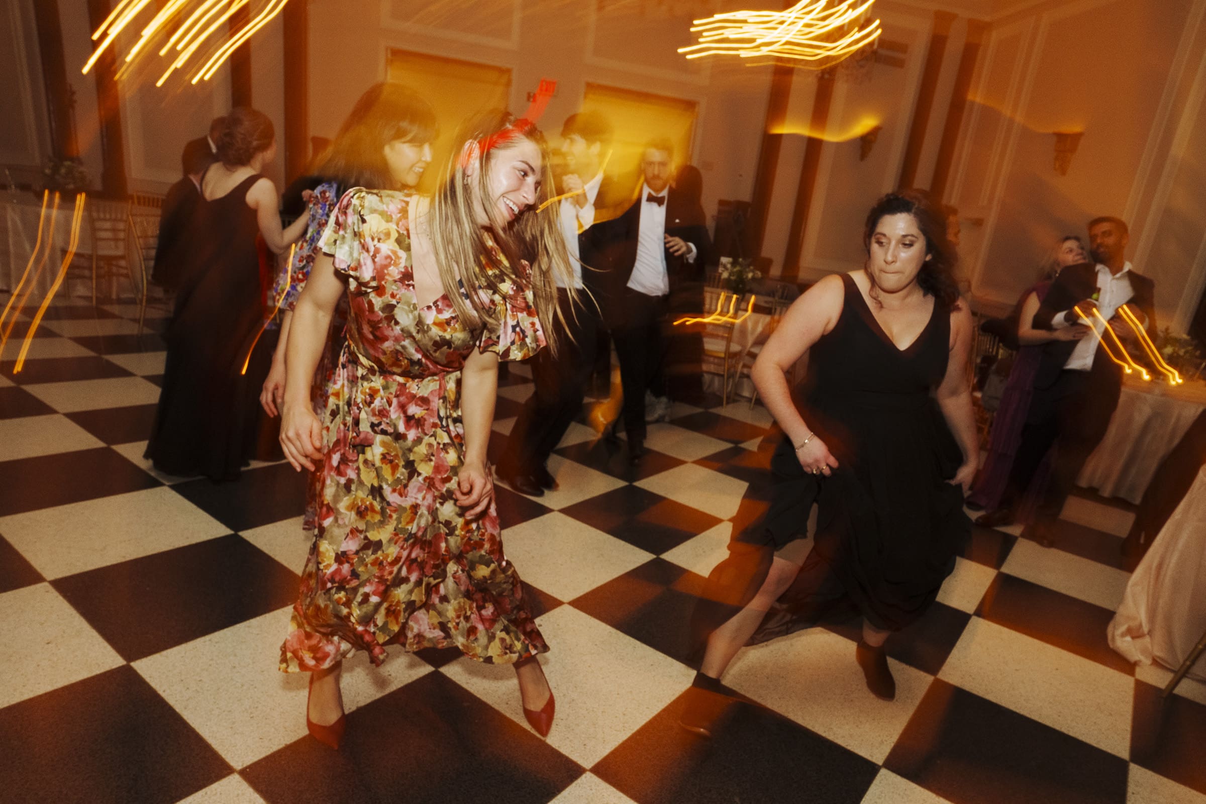 Carolina Inn Wedding - Frances & Lewis In the dimly lit room of the Carolina Inn wedding, guests are joyfully dancing on a black and white checkered floor. The lively atmosphere is captured with motion blur. A woman in a floral dress and another in a black dress take center stage in the vibrant scene.