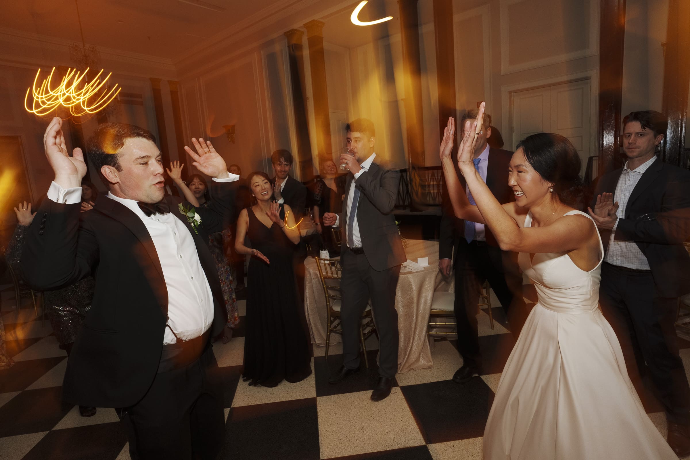 Carolina Inn Wedding - Frances & Lewis A joyful wedding reception at the Carolina Inn with guests dancing energetically under warm lighting. A bride in a white dress and a groom in a tuxedo lead the celebration on a checkered floor, surrounded by elegantly dressed friends.
