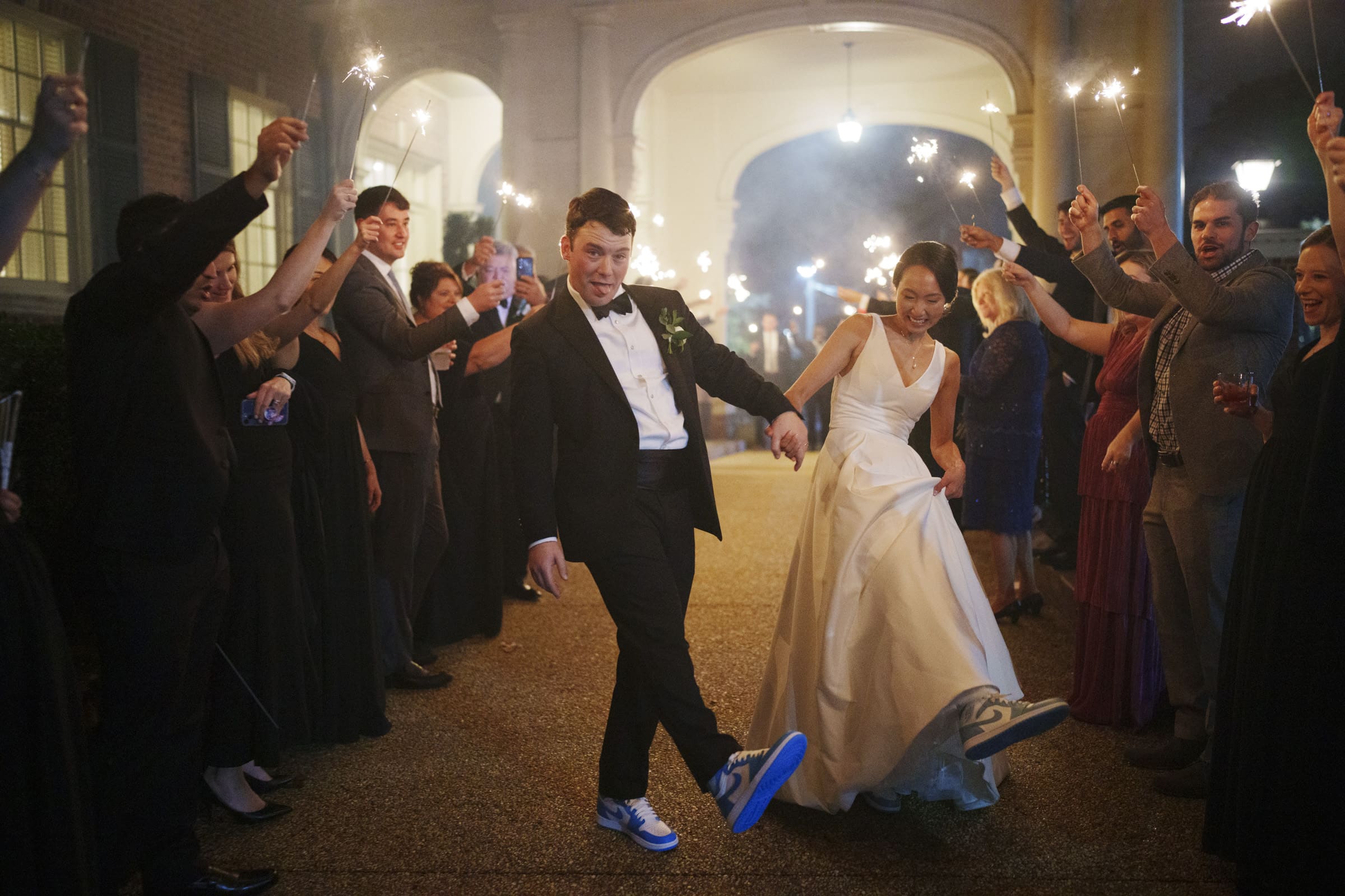 Carolina Inn Wedding - Frances & Lewis A joyful bride and groom in formal attire walk under an archway at their Carolina Inn wedding. The bride sports a white dress with sneakers, while the groom complements her style with a tuxedo and sneakers. Guests lining the path hold sparklers, celebrating their sparkling exit.