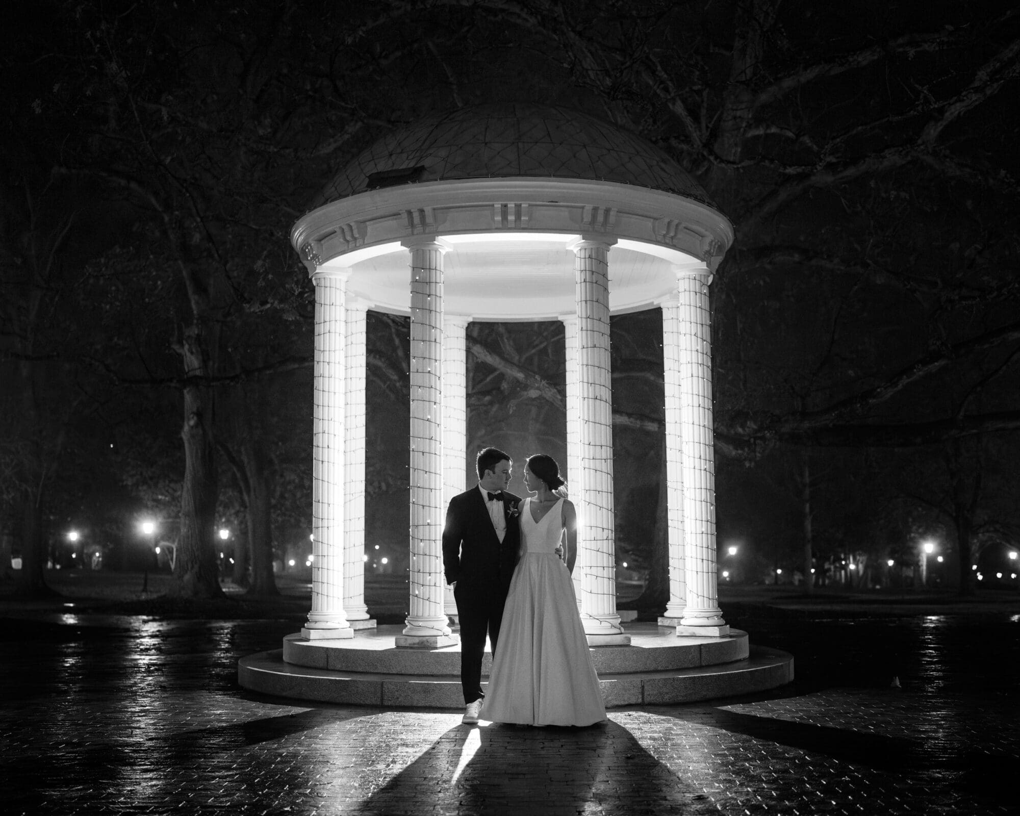Carolina Inn Wedding - Frances & Lewis A bride and groom stand under an illuminated gazebo at night during their Carolina Inn wedding. The bride wears a long dress, and the groom is in a suit. The black-and-white scene features trees and soft lights in the background, creating a romantic atmosphere.