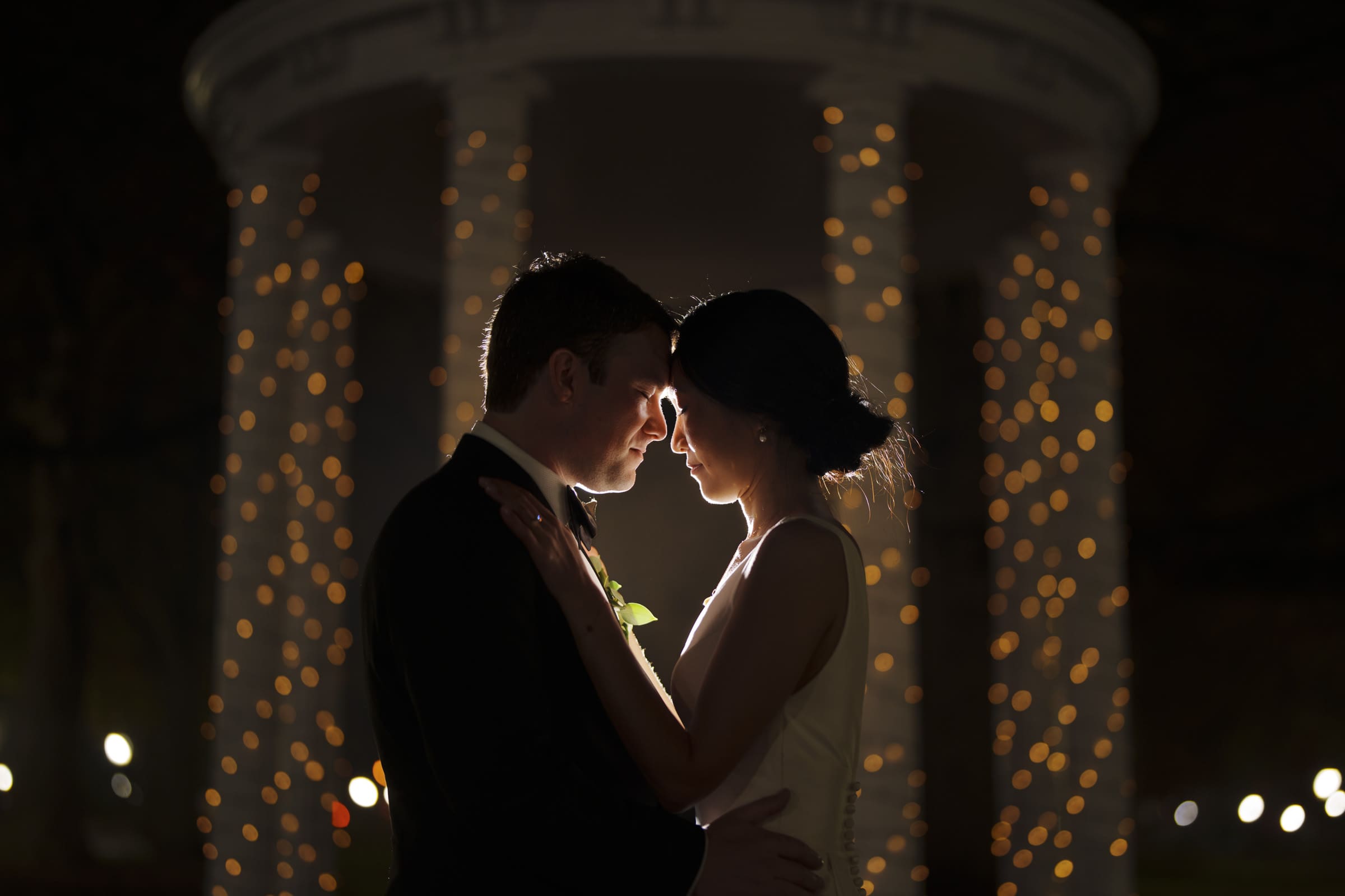 Carolina Inn Wedding - Frances & Lewis At a Carolina Inn wedding, a couple in formal attire stands close, facing each other under a softly lit gazebo adorned with string lights. The warm glow creates an enchanting, romantic atmosphere that perfectly captures the elegance of their special night.