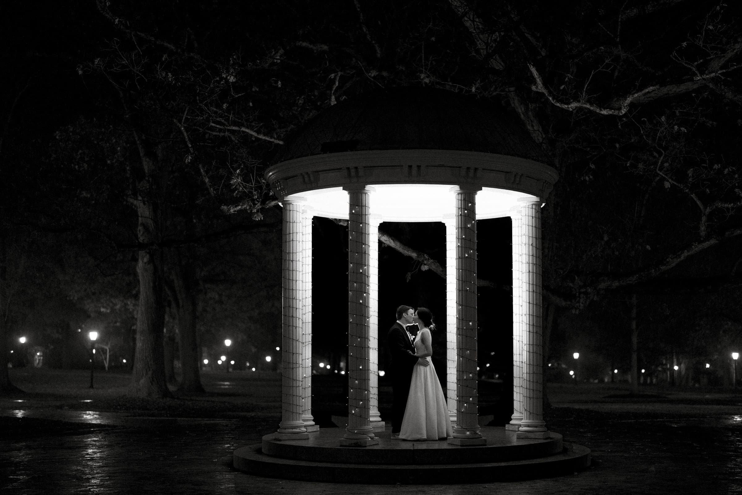 Carolina Inn Wedding - Frances & Lewis A couple stands under a lit gazebo at night, surrounded by darkness and trees. They embrace each other in a romantic setting, reminiscent of a Carolina Inn Wedding, creating a contrast between the illuminated structure and the shadowy background.