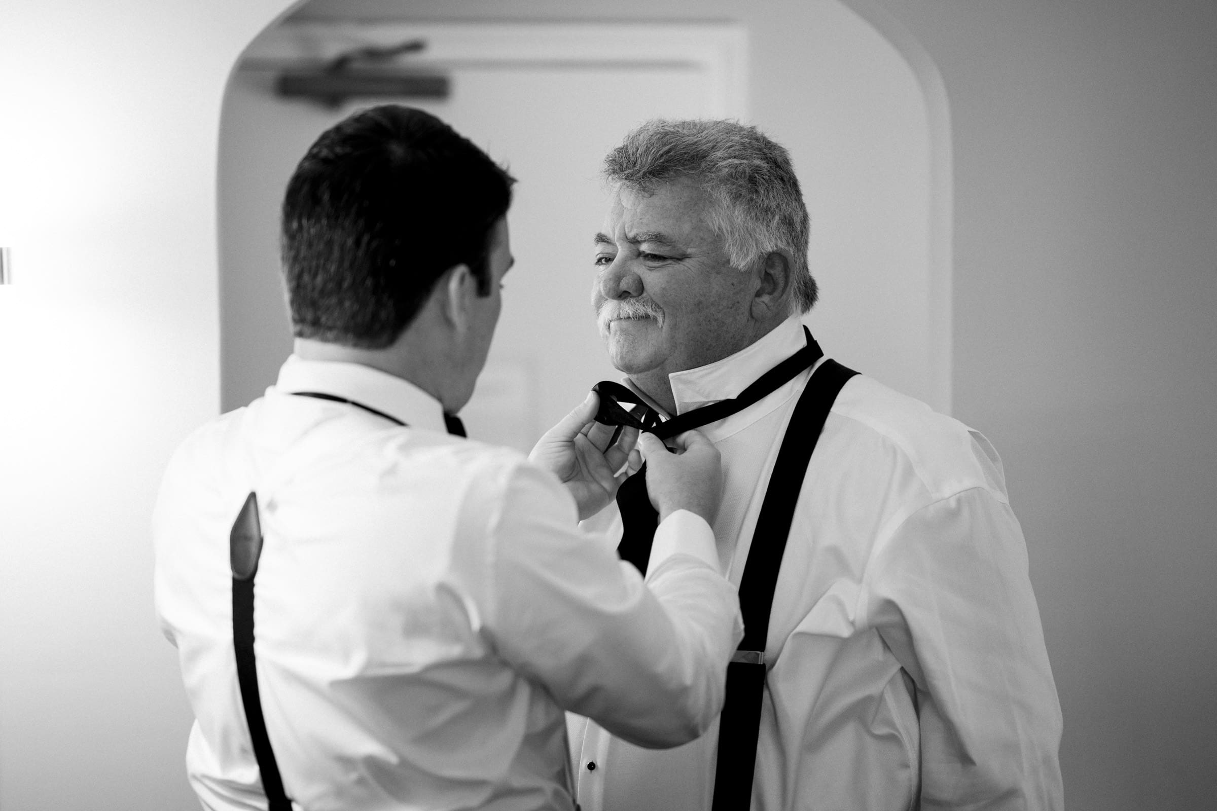 Carolina Inn Wedding - Frances & Lewis Two men in dress shirts and suspenders are in a room at the Carolina Inn. One is helping the other adjust his bow tie. Both appear to be in a calm, focused moment. The image is in black and white, creating a classic, timeless feel perfect for a wedding day preparation.