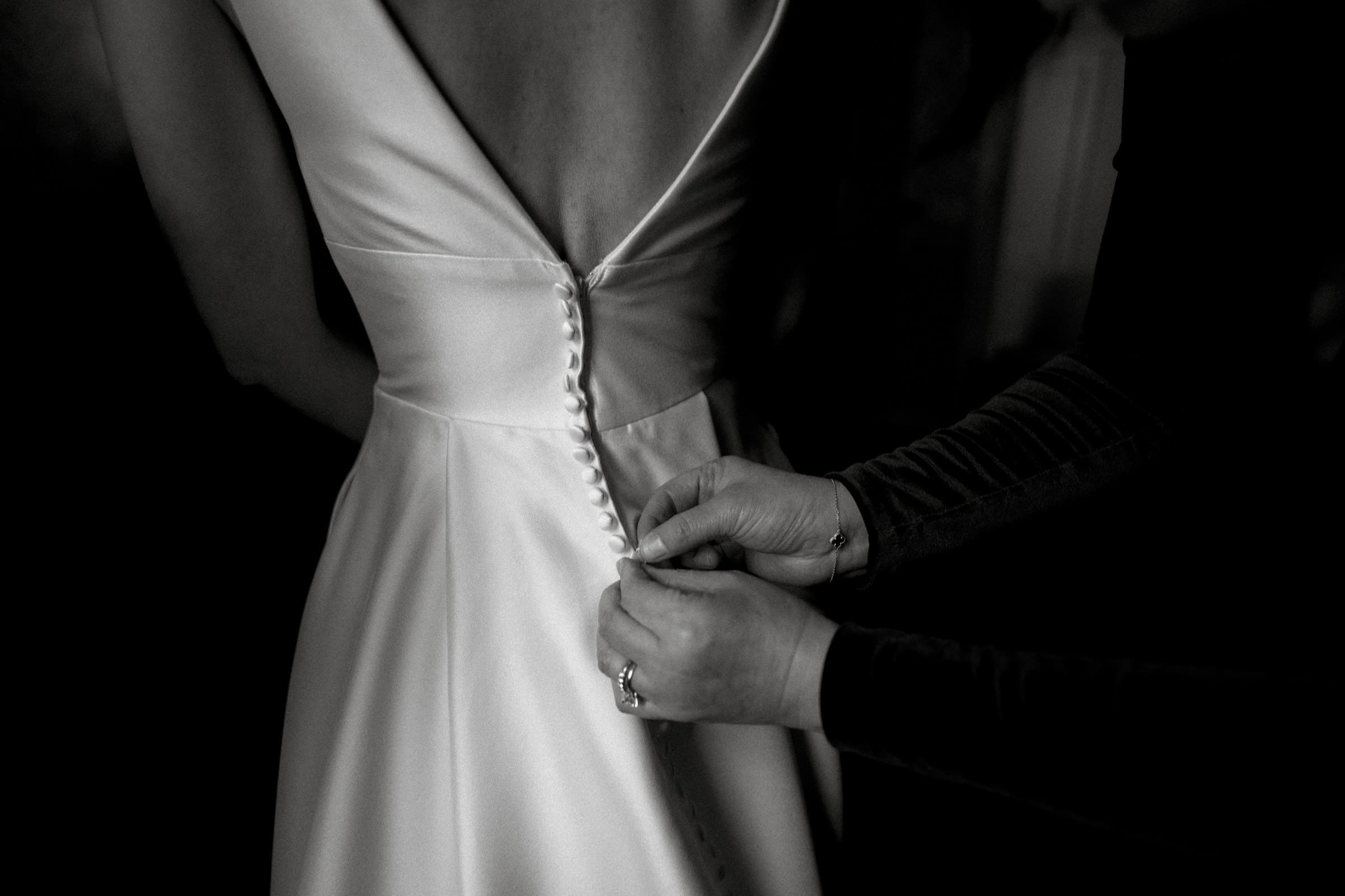 Carolina Inn Wedding - Frances & Lewis A close-up of a person fastening the buttons on the back of a wedding dress at a Carolina Inn Wedding. The elegant dress features a deep V-back, and the hands gently securing the buttons create an intimate and timeless scene in black and white.