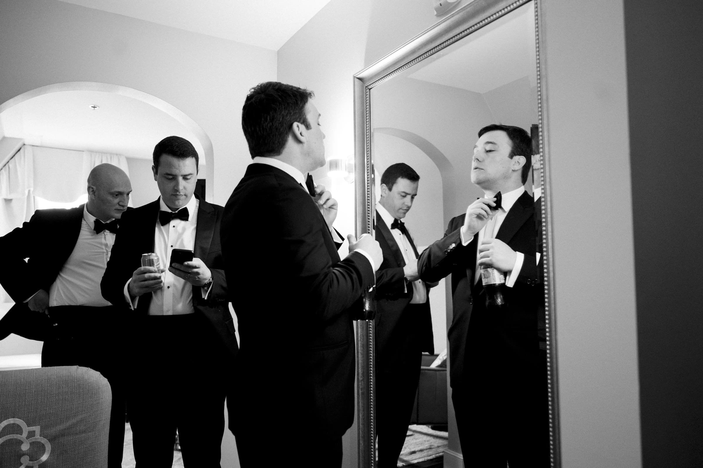 Carolina Inn Wedding - Frances & Lewis A group of men in tuxedos are gathered in a room at the Carolina Inn. One man adjusts his tie in front of a large mirror, while another checks his phone. A third holds a drink. The elegant decor complements the formal yet relaxed atmosphere, setting the perfect tone for the wedding preparations.