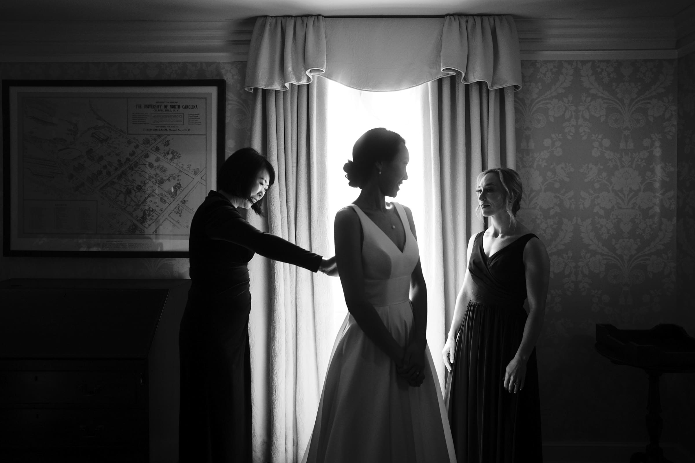 Carolina Inn Wedding - Frances & Lewis A bride in a white dress stands near a bright window at the Carolina Inn, attended by two women. One adjusts her dress while the other looks on. The room's patterned wallpaper and framed map create an elegant and intimate setting befitting a perfect wedding day.