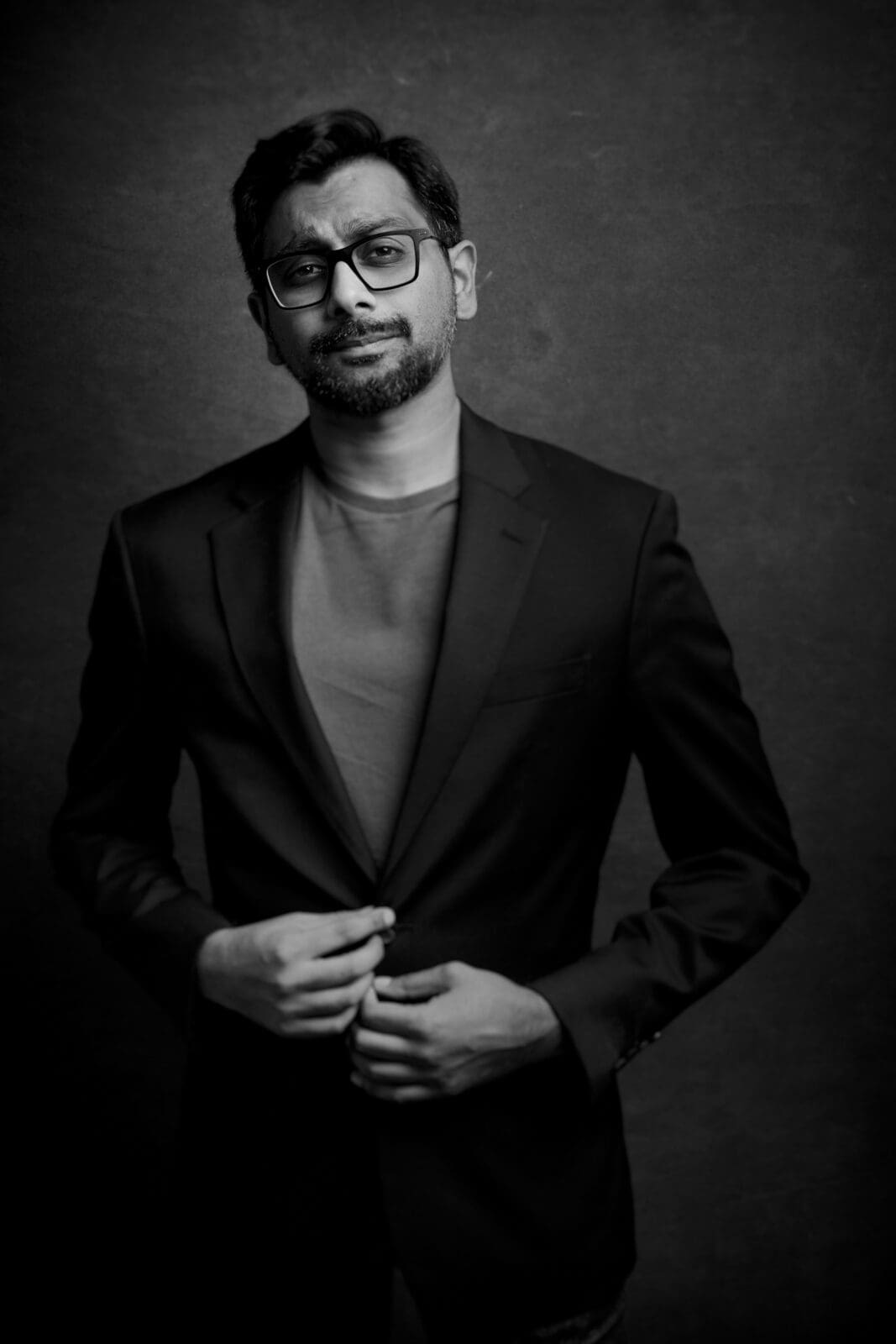 Headshot & branding session for dj rang A black and white headshot captures a man with glasses, wearing a blazer over a t-shirt. With a confident expression, he adjusts his jacket against a plain backdrop, embodying the essence of a professional branding session.