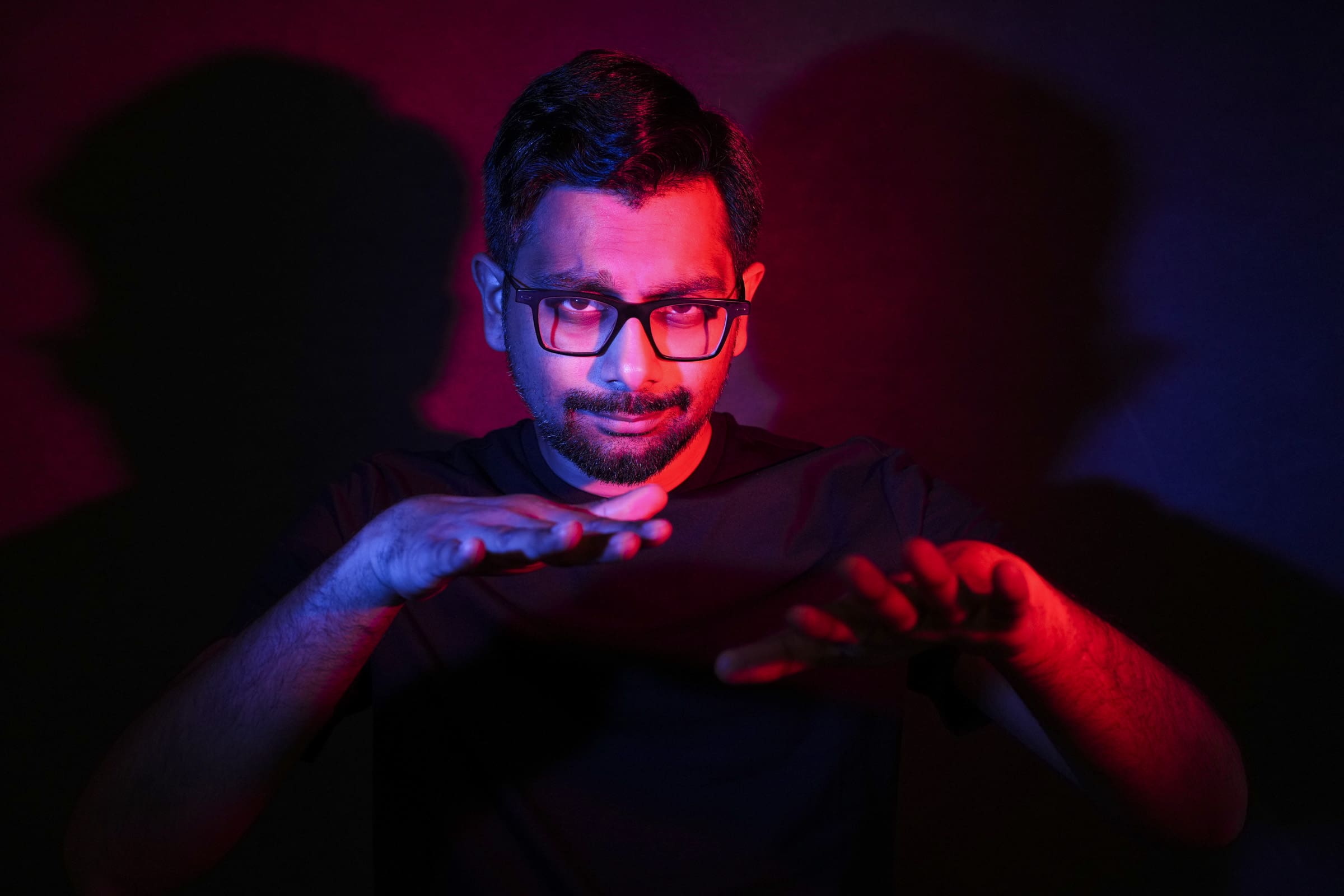 Headshot & branding session for dj rang In a striking headshot, a person with glasses stands against a dark background, dramatically lit by red and blue lights. During this unique branding session, their raised hands cast intriguing shadows on the wall behind them.