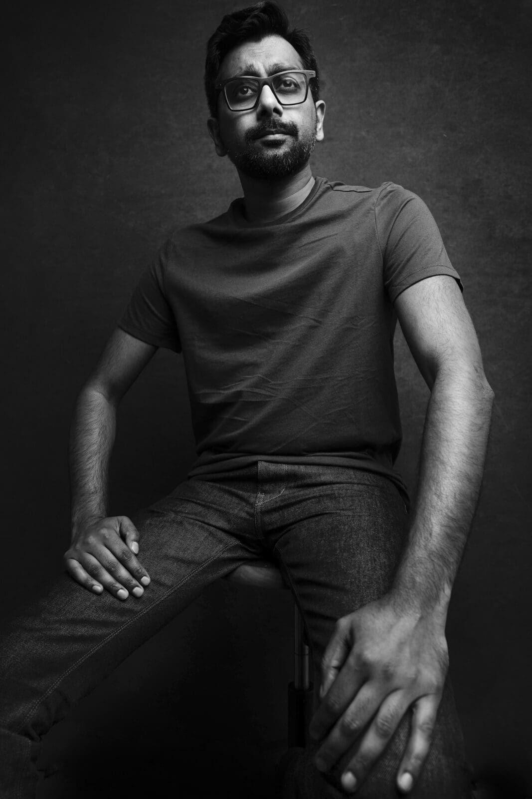 Headshot & branding session for dj rang A black and white headshot captures a person with glasses, wearing a plain t-shirt and jeans, seated against a dark textured background. The thoughtful expression enhances the branding session's tone, with hands resting gently on their legs.
