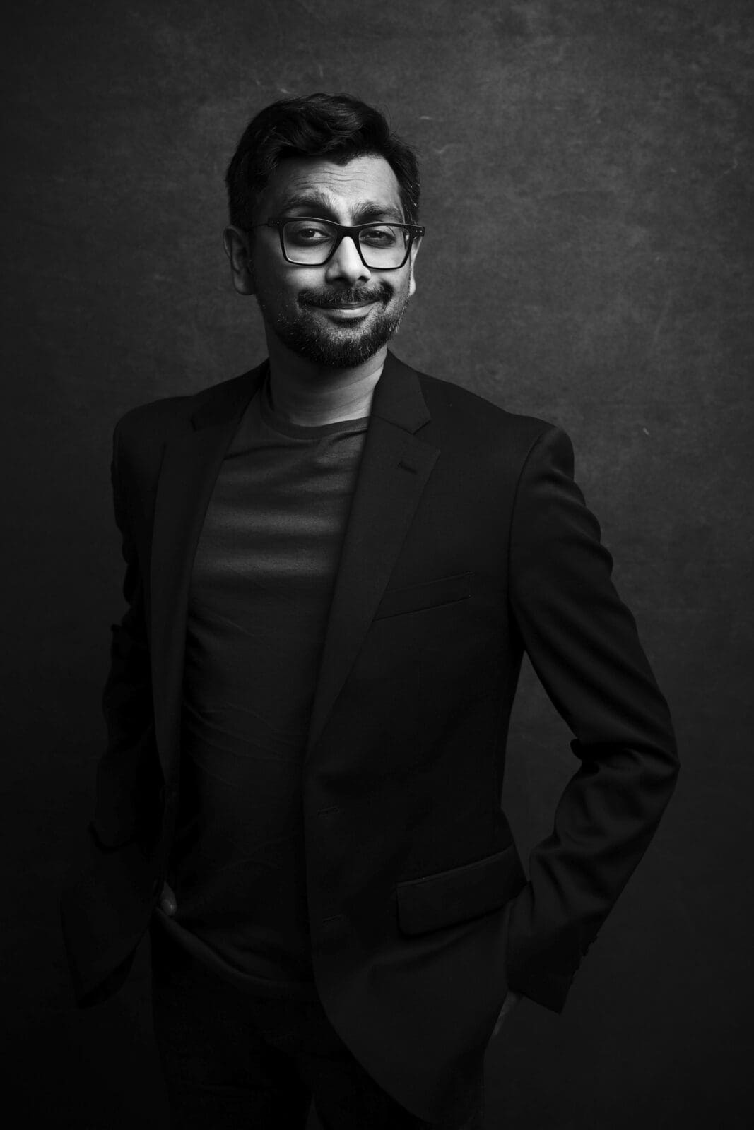 Headshot & branding session for dj rang A black and white headshot of a man wearing glasses and a suit jacket. He is smiling slightly with his hands in his pockets against a dark background, capturing the essence of a refined branding session.