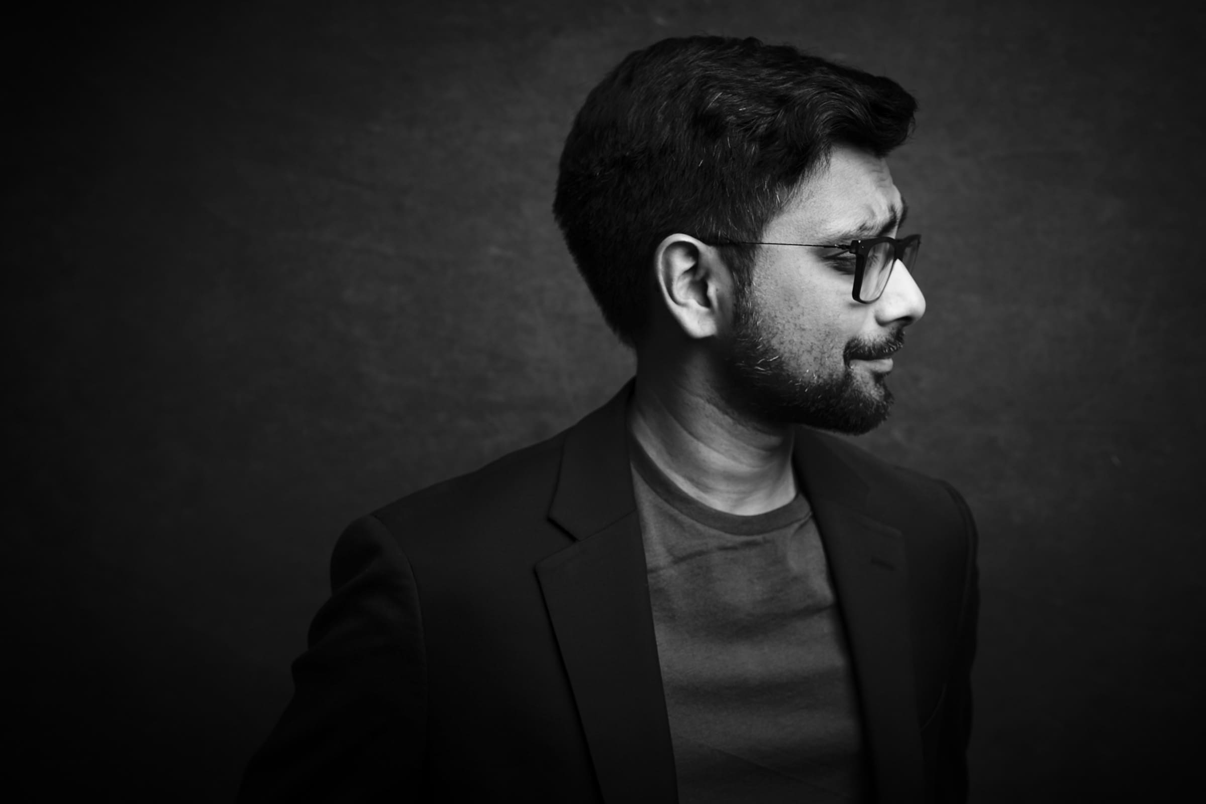 Headshot & branding session for dj rang Black and white headshot of a man with glasses, a beard, and short hair, wearing a blazer and t-shirt. Captured in a branding session against a dark background, he is facing sideways.