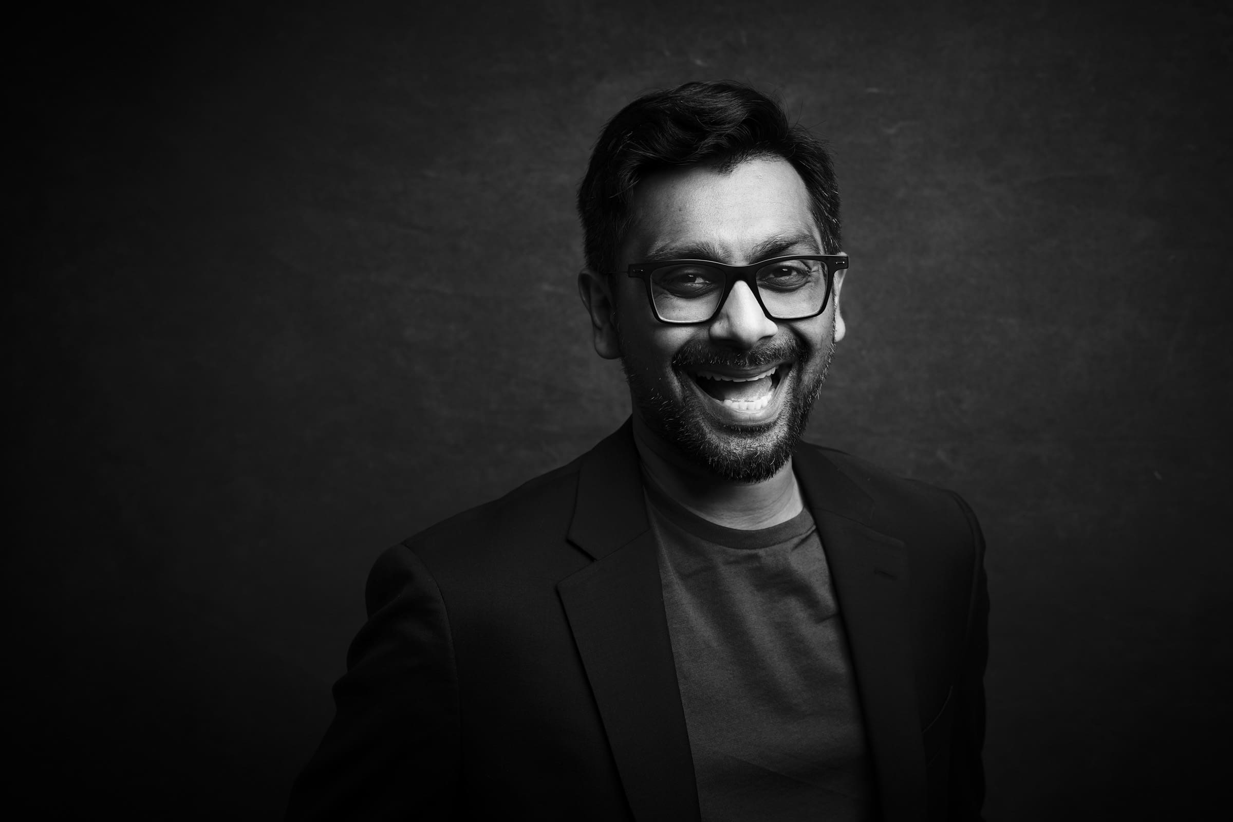 Headshot & branding session for dj rang Black and white headshot of a smiling man wearing glasses, a dark jacket, and a t-shirt. Captured during a branding session, he is looking at the camera against a plain dark background.